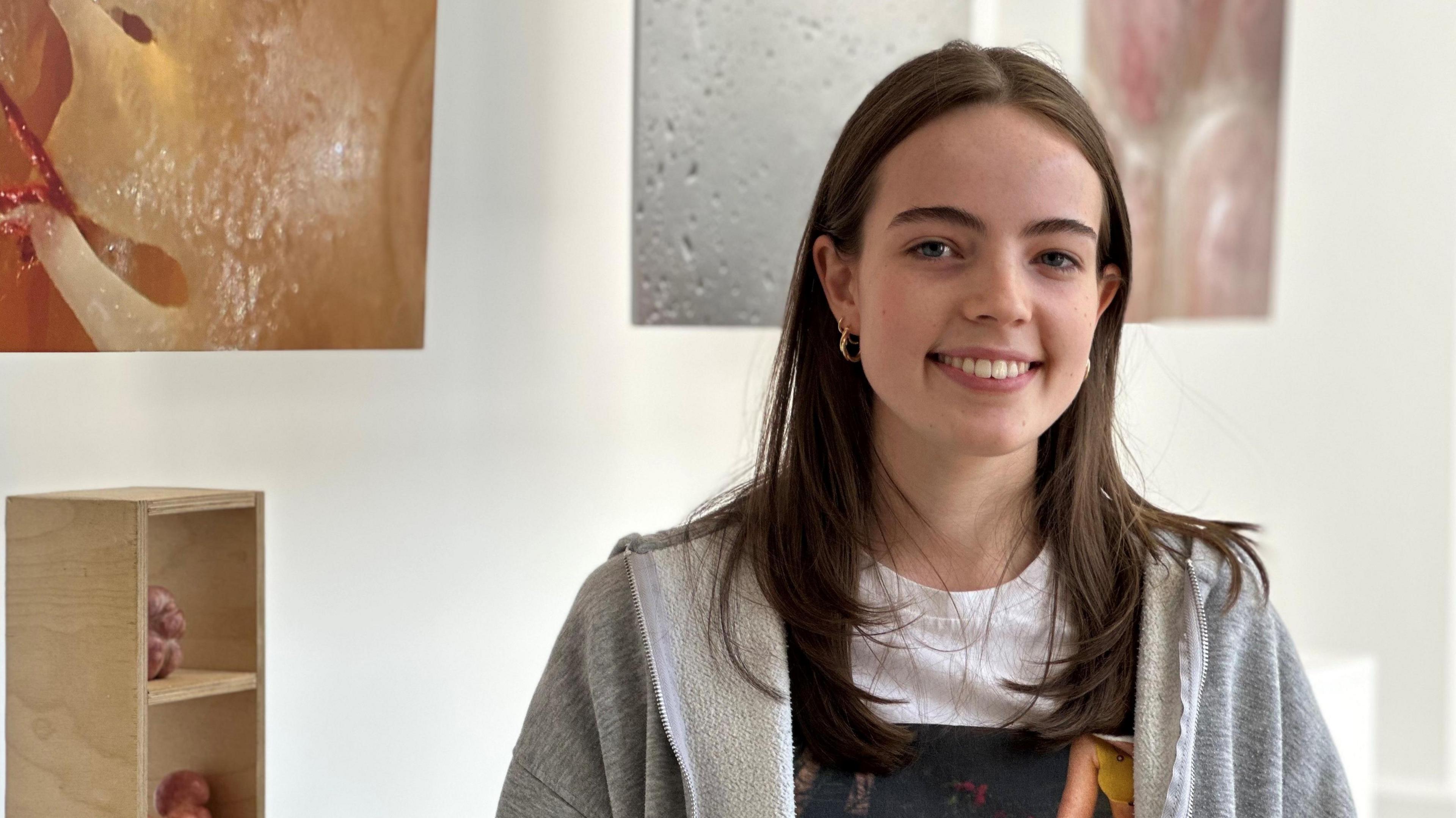 Lula Thomas has long brown hair wearing a white and grey top. Her art work is hanging behind her.