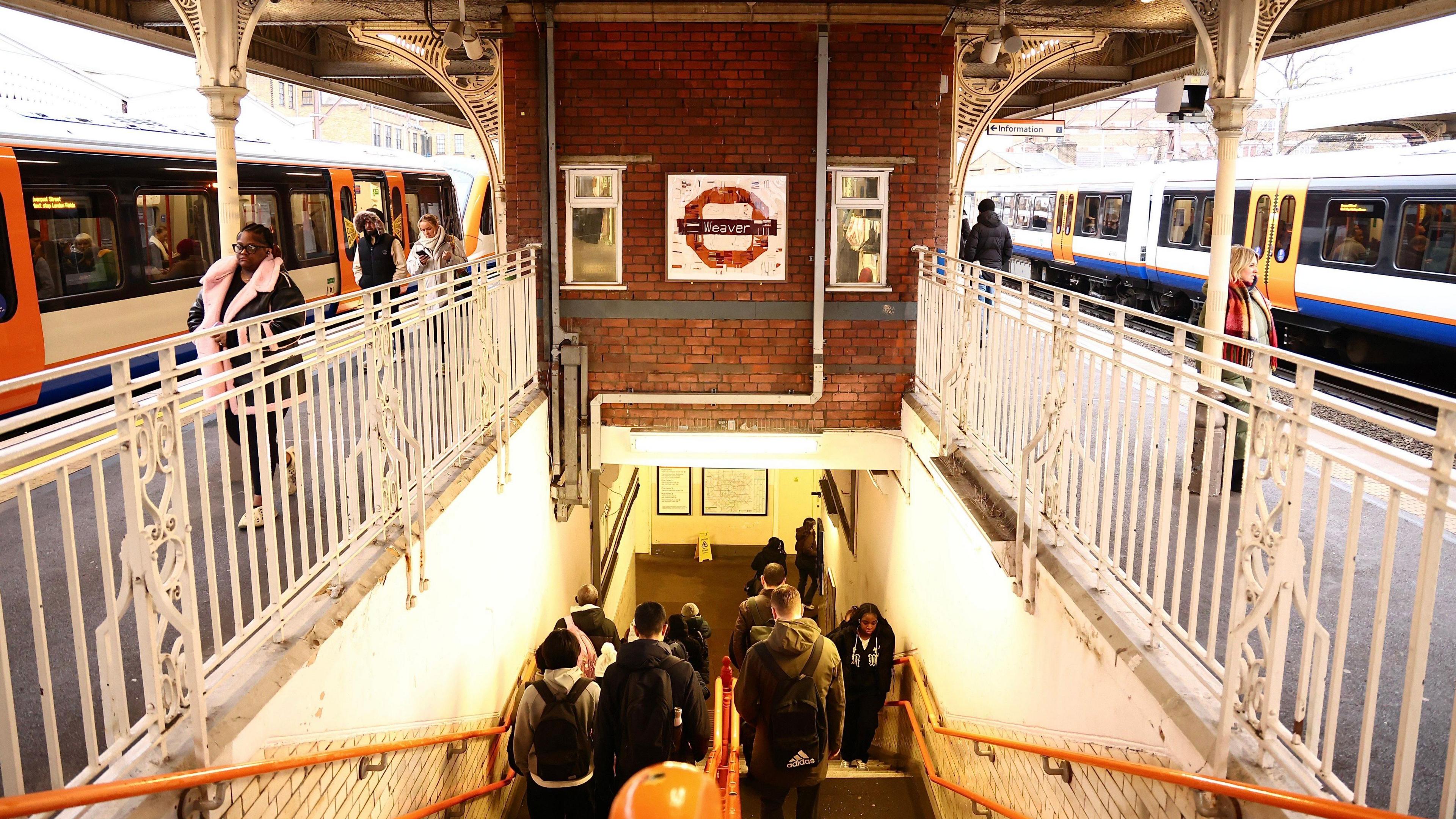 A London Overground station scene at Hackney Downs, featuring the Weaver line roundel mounted on a red brick wall above the stairwell, with passengers walking between platforms and boarding trains