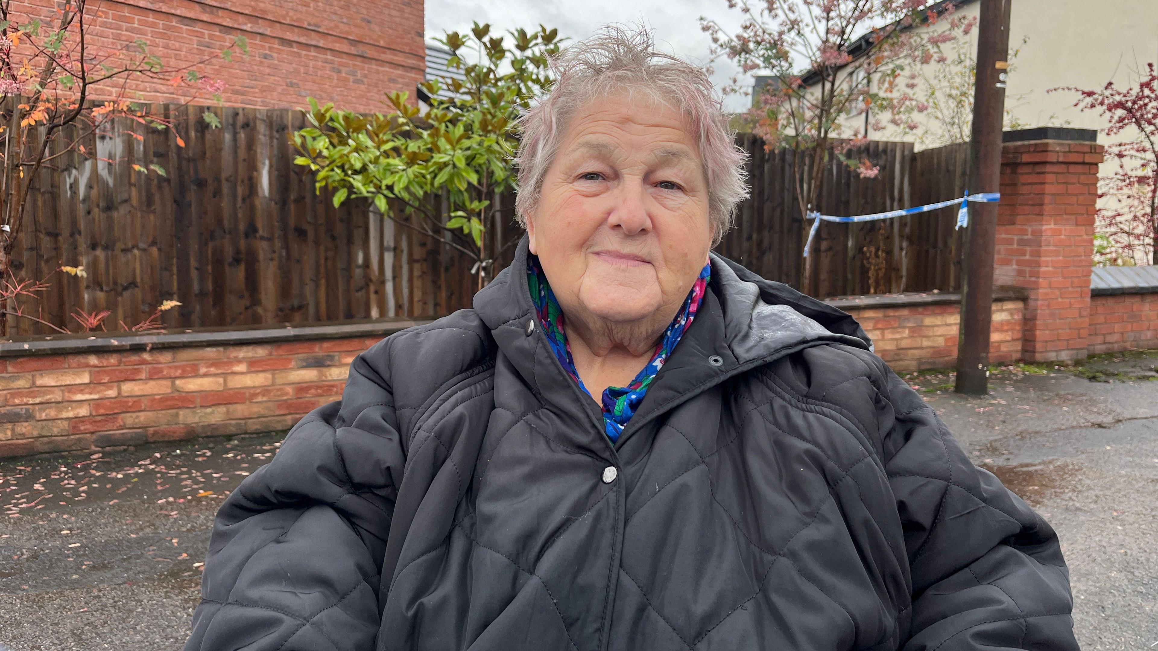 A woman with short grey and pink hair, wearing a blue, green and pink top with a black quilted jacket over the top. She is standing on a pavement that is wet from rain, and there is a wooden fence behind her, as well as blue and white police tape.