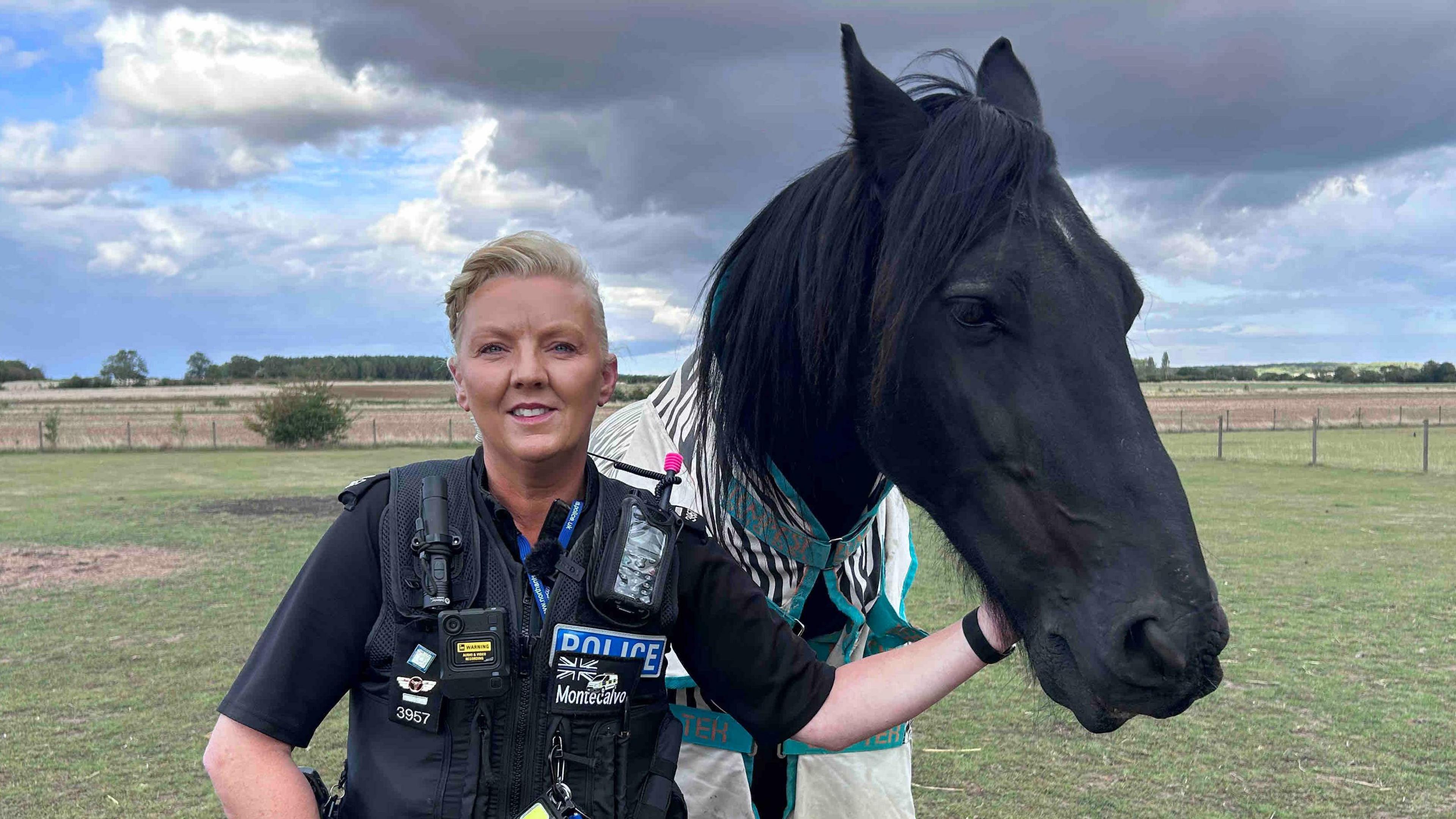Lorna Montecalvo standing in a field in her police uniform. She has blonde hair and is looking directly at the camera and smiling. She is stood next to a black horse named Falcon.