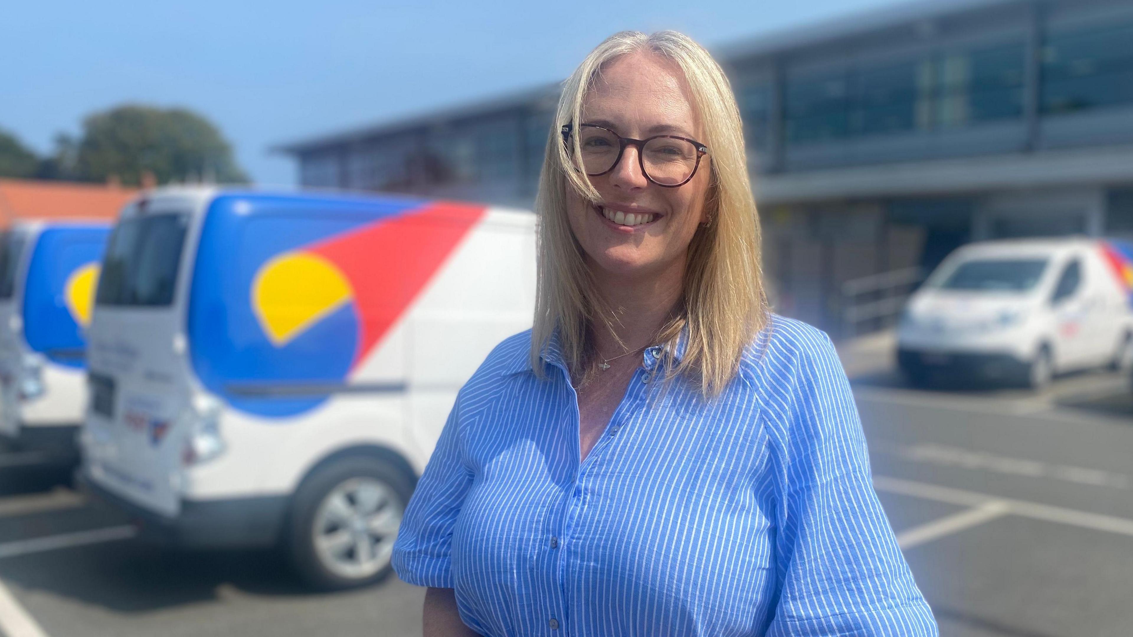 Dawn has shoulder-length straight blonde hair. She is wearing glasses and a blue shirt with white stripes. She is standing outside the Guernsey Post headquarters.