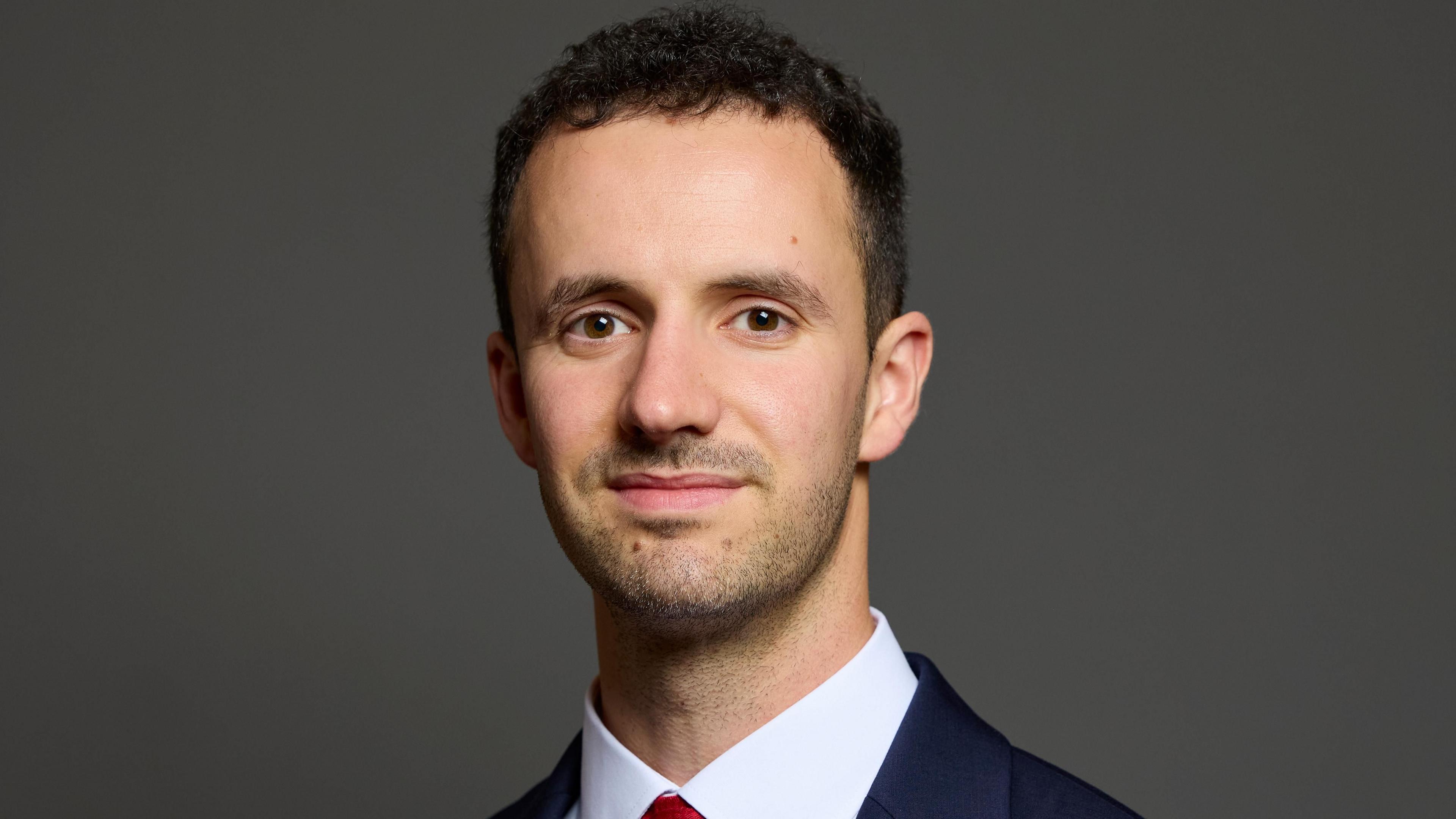 Chris Hinchliff wearing a suit with a white shirt and red tie. He is looking directly at the camera and smiling.
