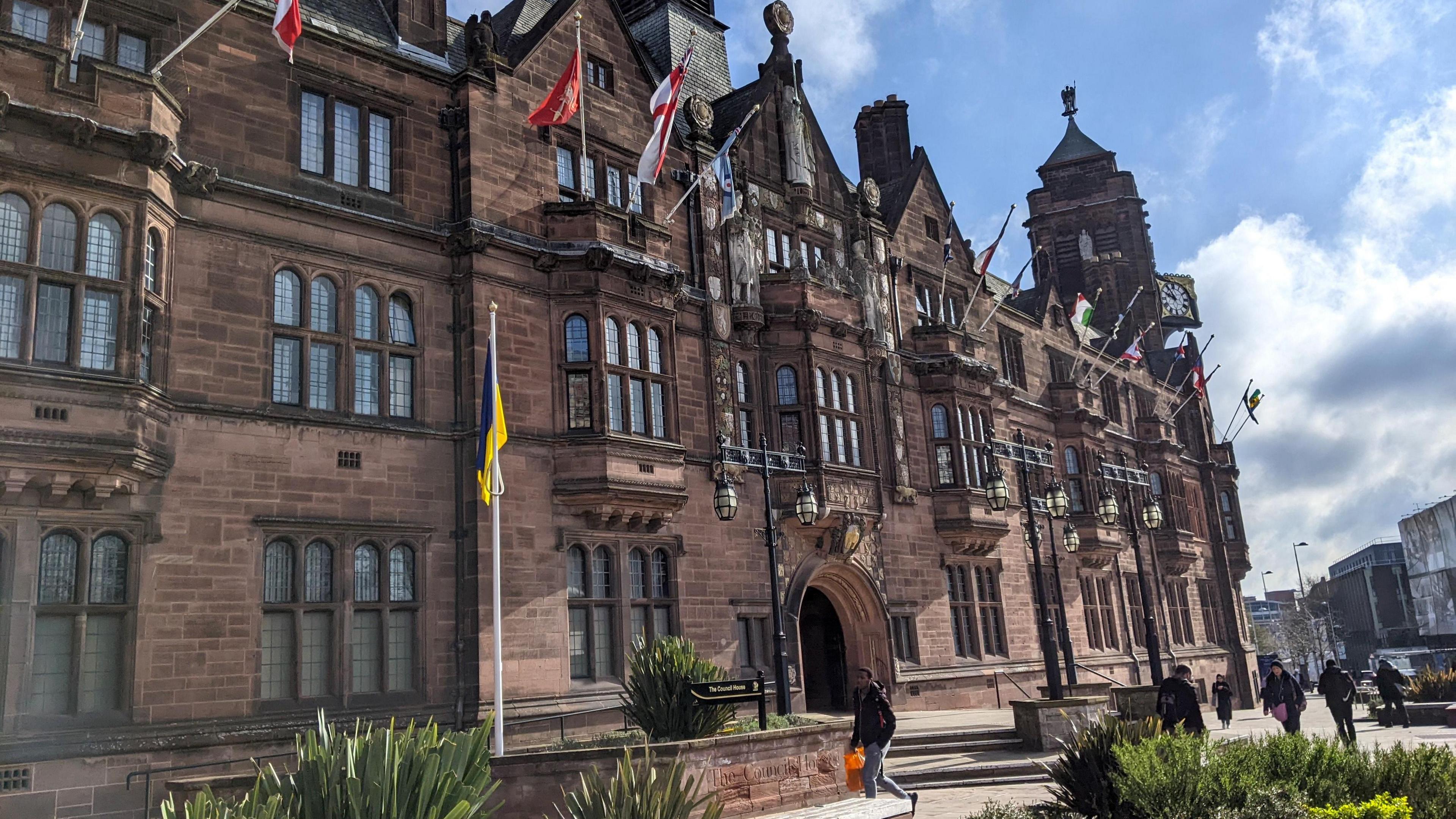 Coventry City Council offices are in a historic building that was constructed in the 20th Century in an early Tudor style. The building has pedestrian areas outside and flower beds and there are flags decorating the building. People are walking around the grounds.