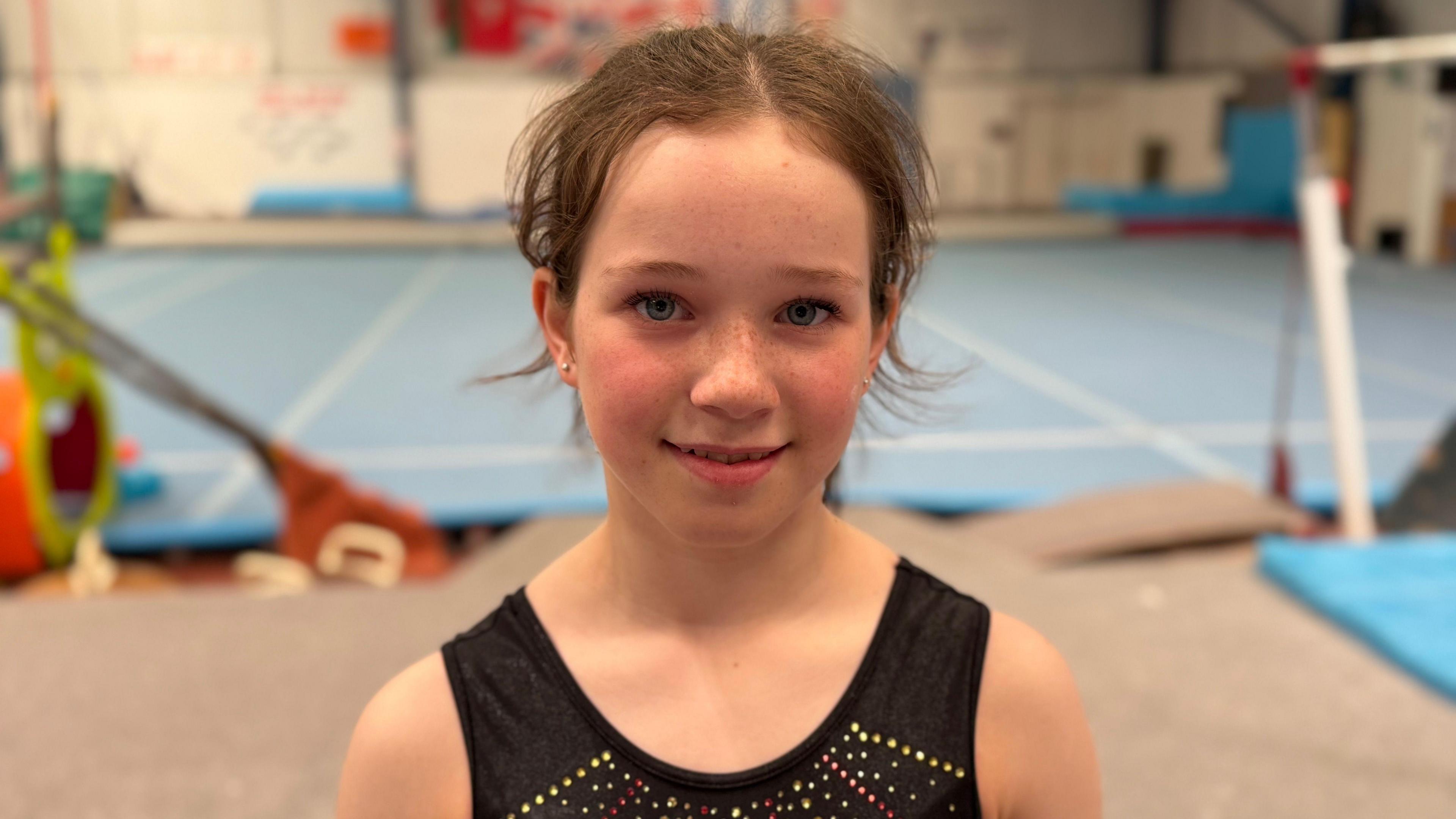 Phoebe Christian smiles, she wears a black vest with yellow, red and white sparkles. Behind her you can see a gymnasium, which has equipment for floorwork and a bar.