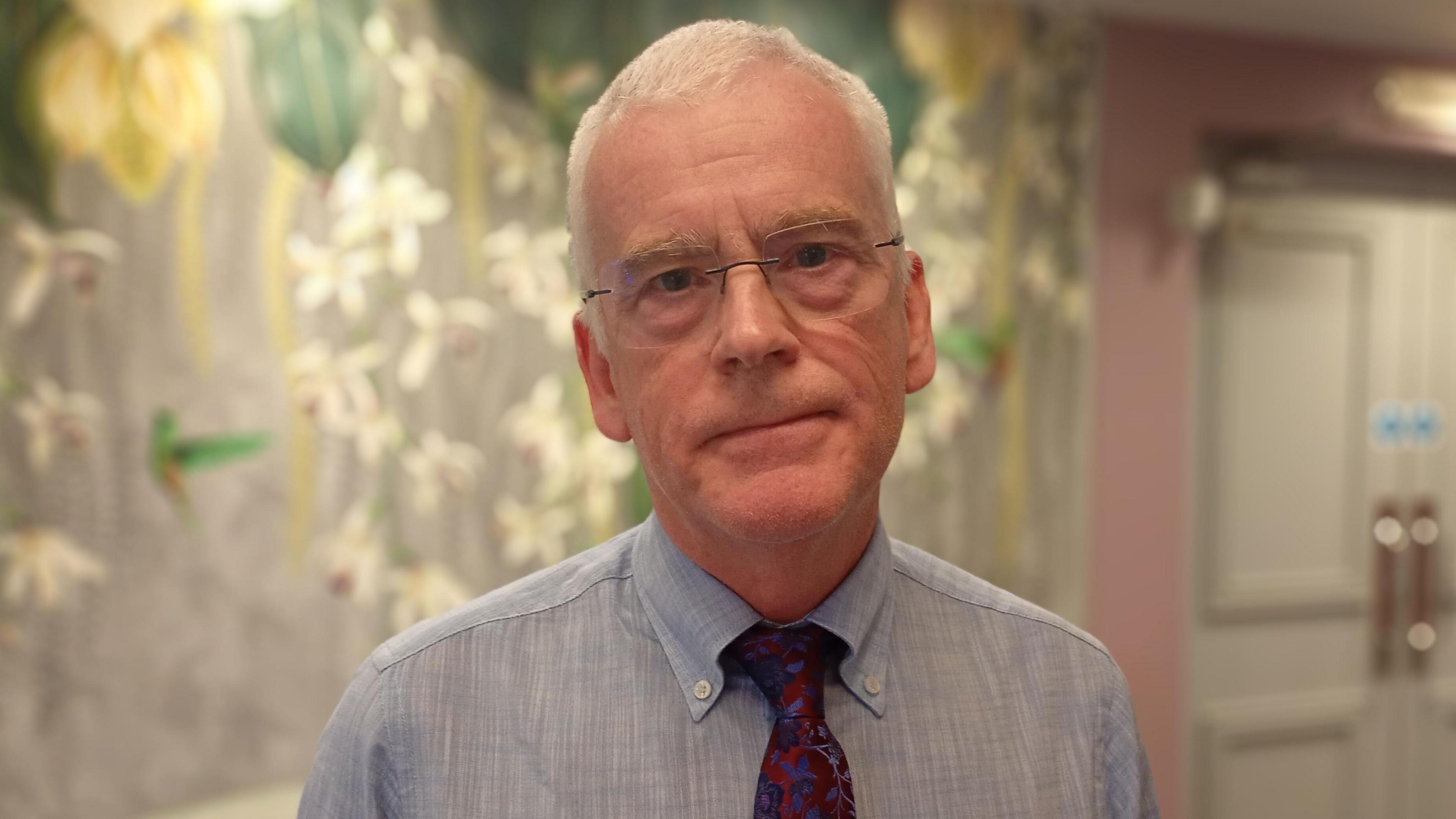 A man, with short white hair, wearing glasses, a dark red tie and a blue shirt.