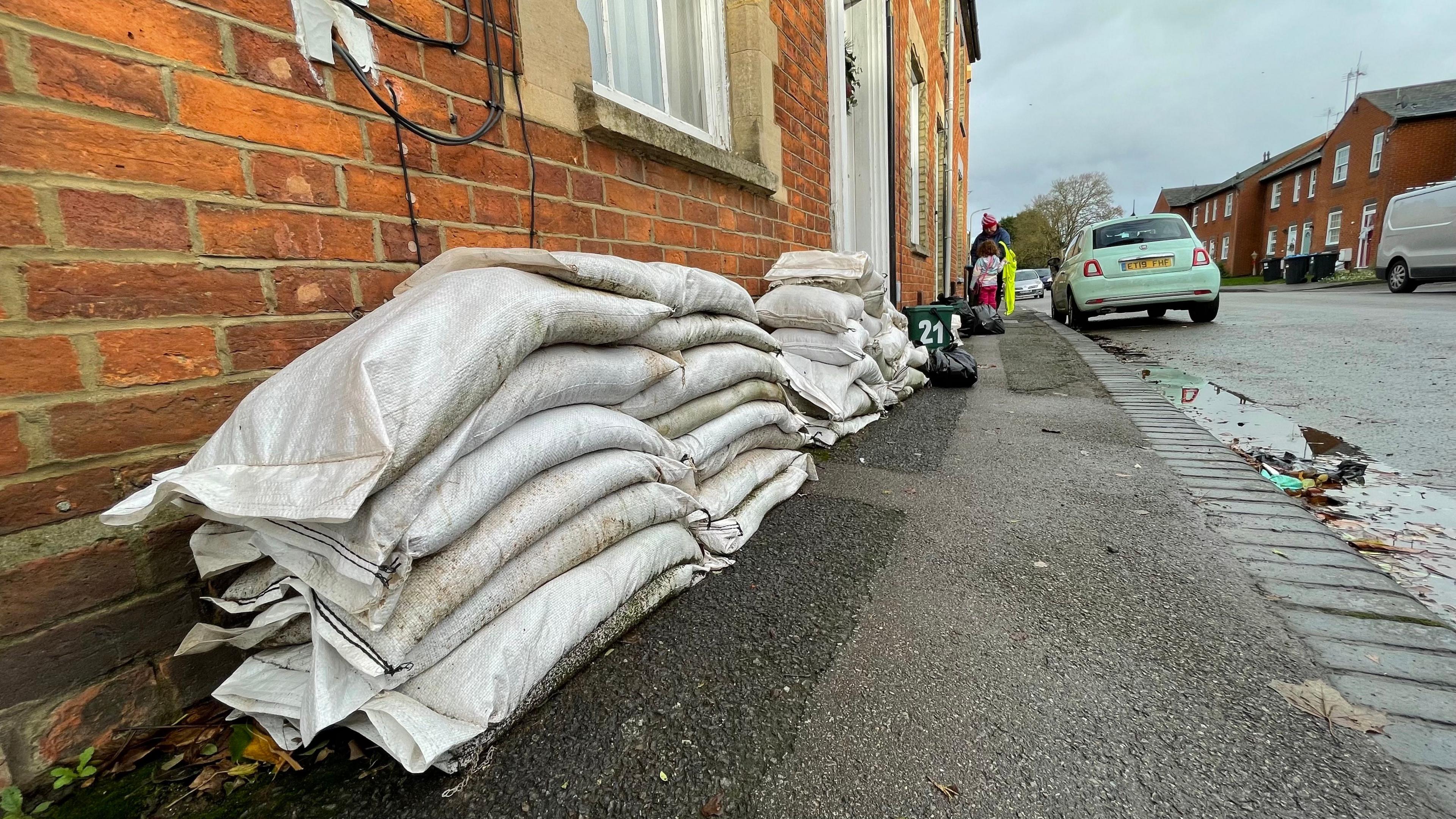 Piles of white sandbags leaning against the fronts of terraced houses in Newport Pagnell. The image is taken looking along the pavement, which disappears into the distance. To the right of the image is the road, with some cars parked up to the sides. The houses are red brick, Victorian-looking terraces.