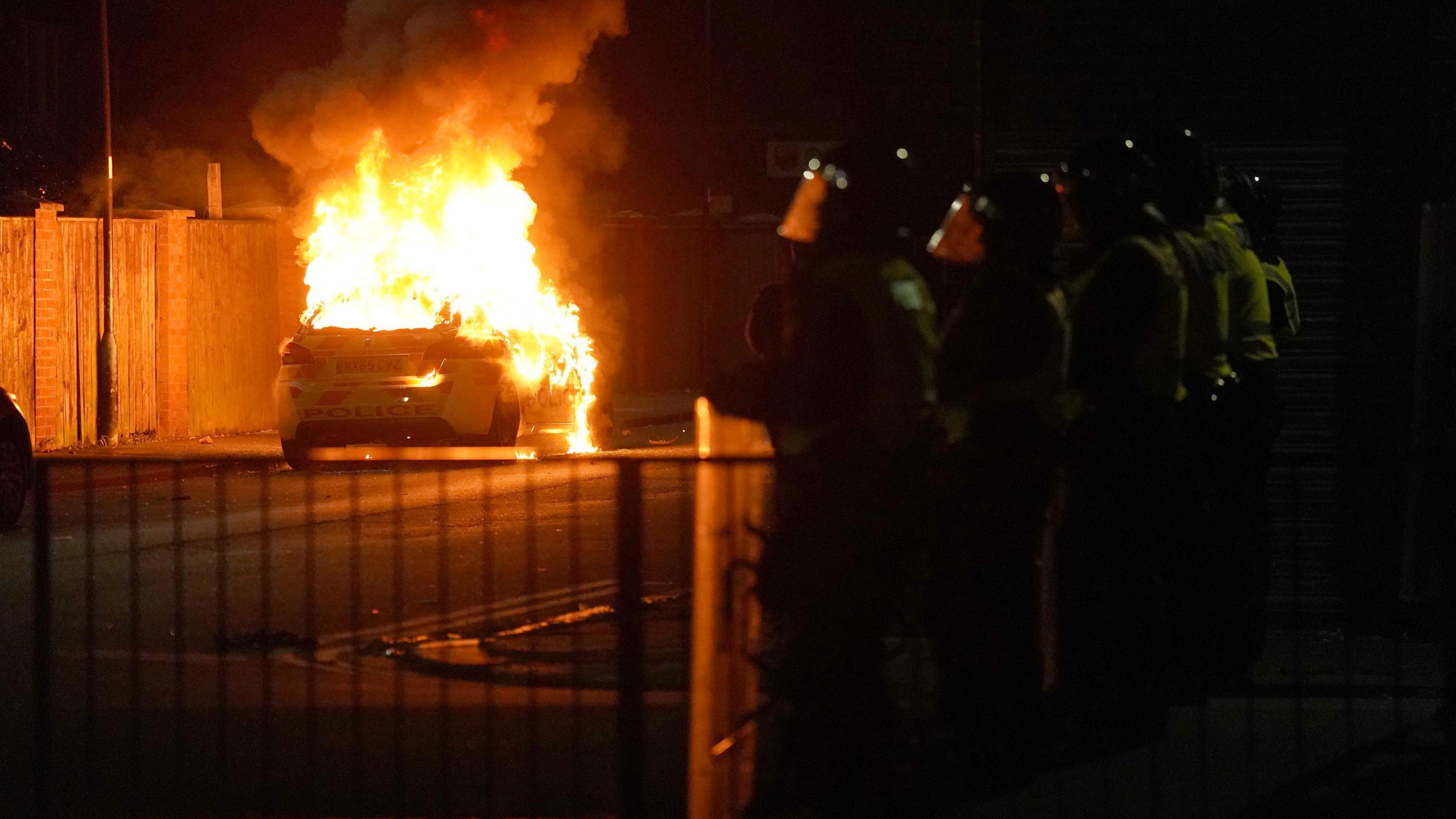 Flames engulfing a police car as police officers in riot gear look on