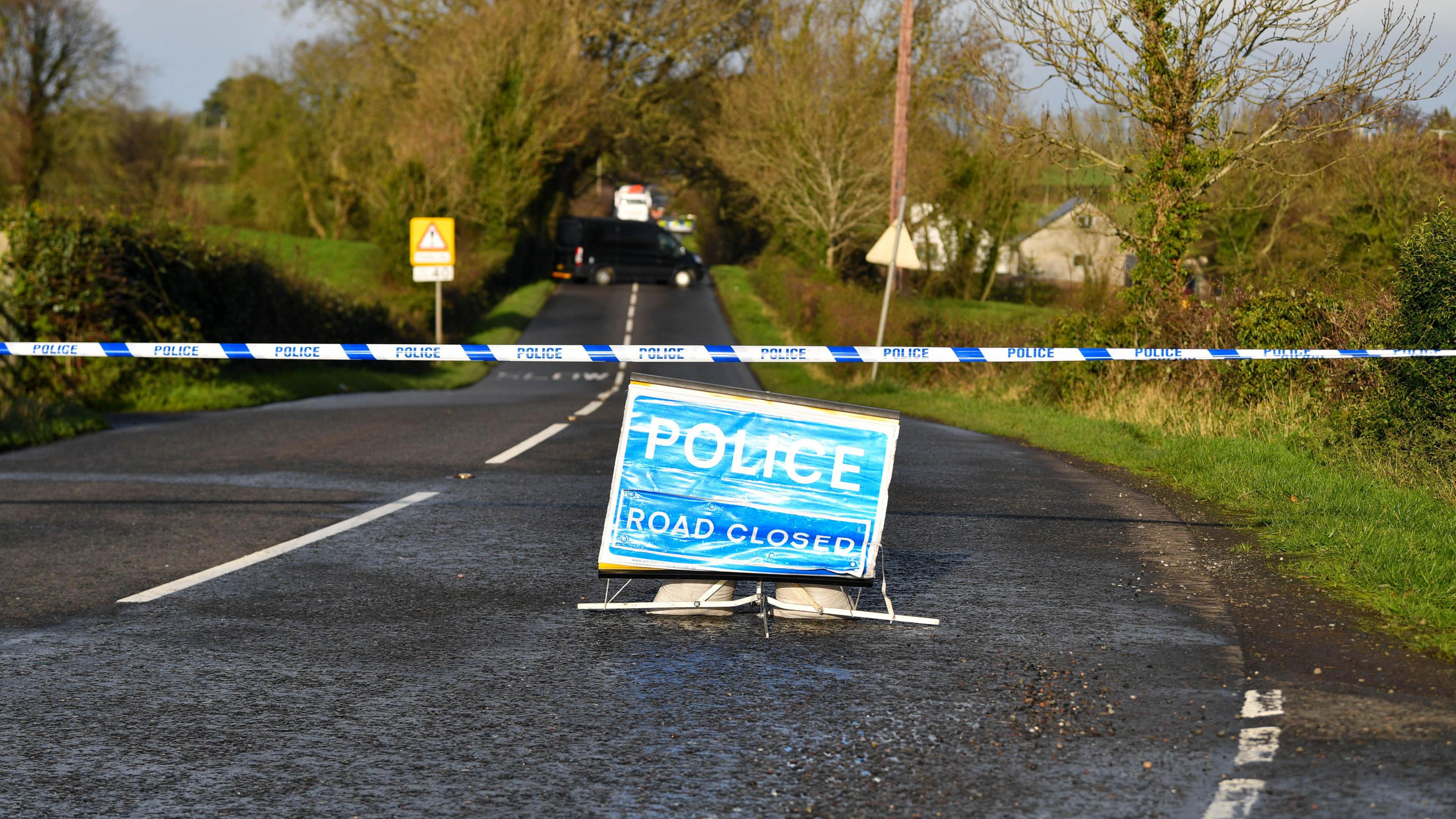 A two lane country road with hedgerows and green fields either side. A road closed police sign is on the road and a barrier is across the road. A back van can be seen at the bottom of the road.