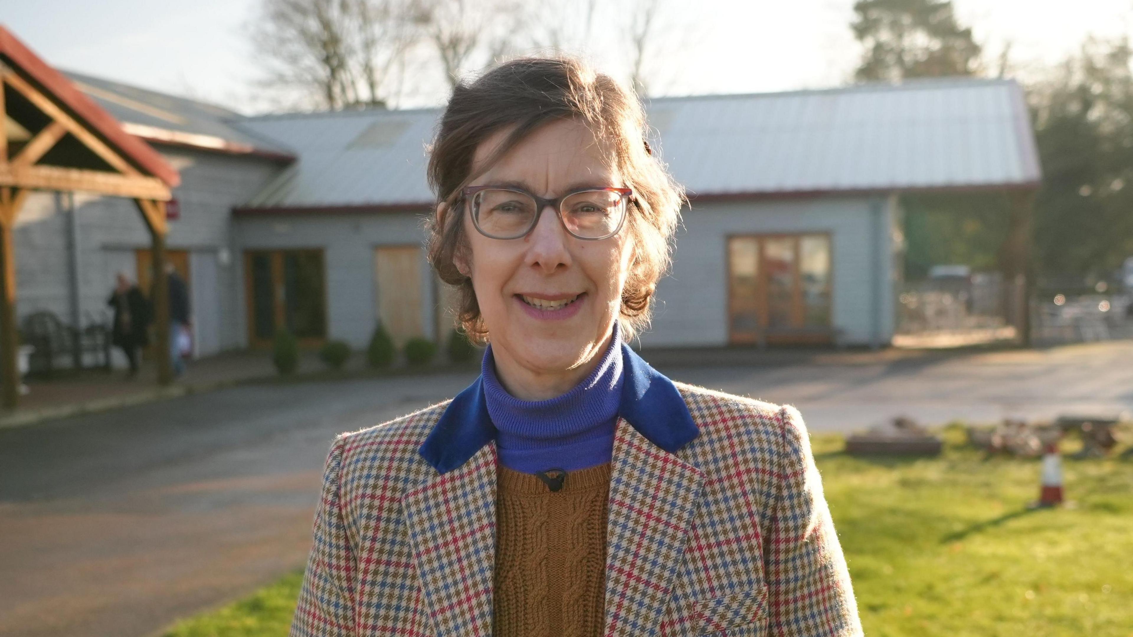 Elizabeth Talbot is looking at the camera and smiling. She is wearing a tweed jacket with a brown jumper and is wearing a pair of glasses. She is stood outside the auction house.