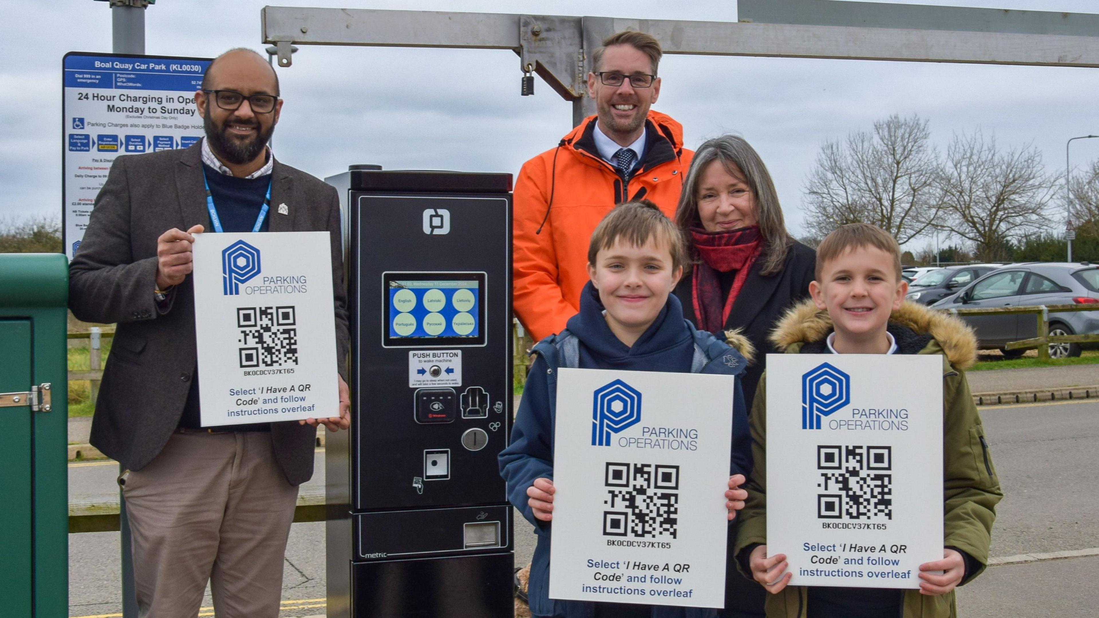 Councillor Bal Anota, head teacher Mat Tuckwood and councillor Deborah Heneghan stand next to the car park pay machine. Bal is wearing a brown suit jacket and cream trousers. Mat is wearing a bright orange coat. Deborah has a red scarf and black coat. 