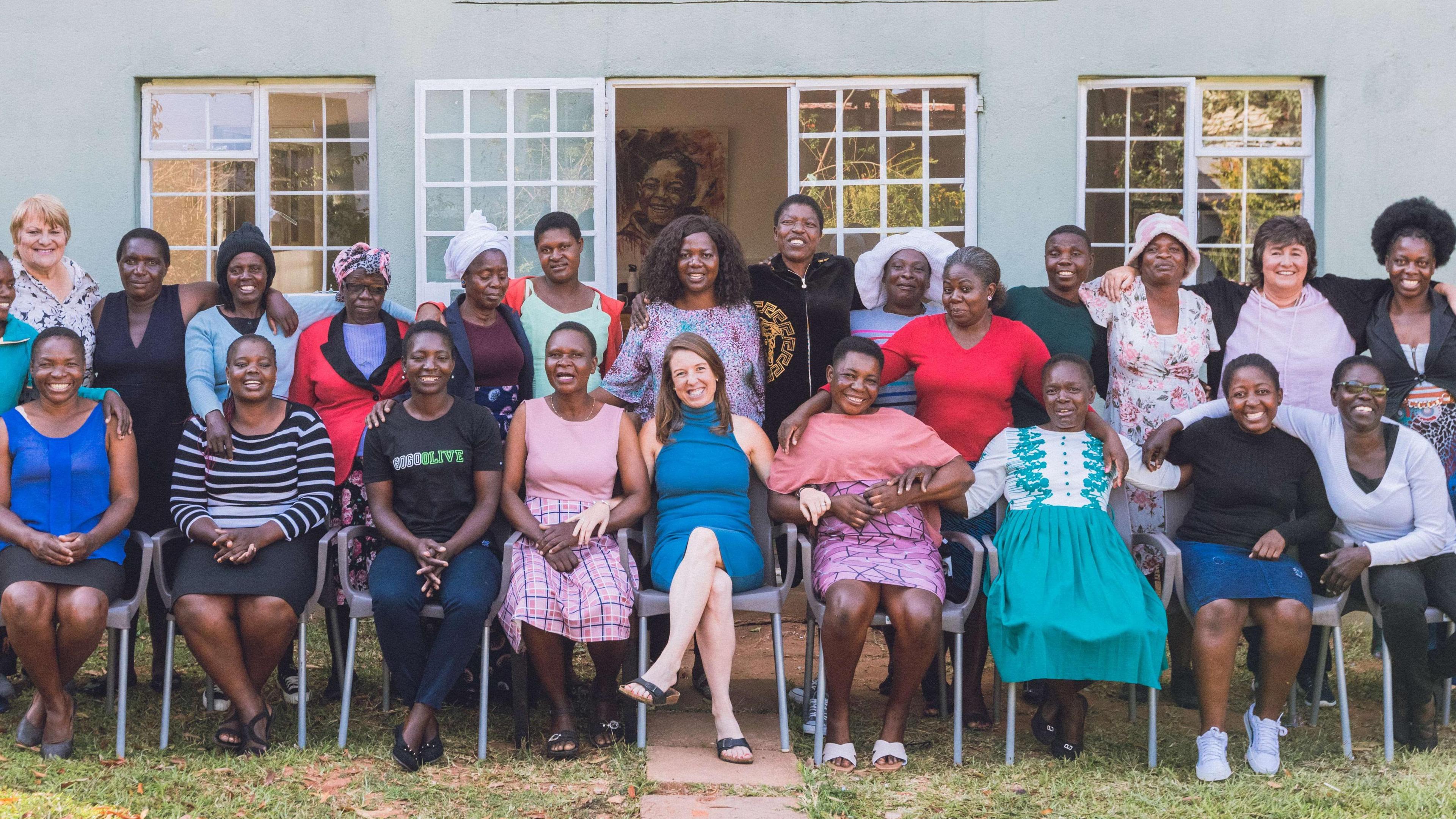 Julie Hagan, in a blue dress and black sandals, sits in the middle of row of smiling women. There is another row of women behind them, standing in front of a grey building. 