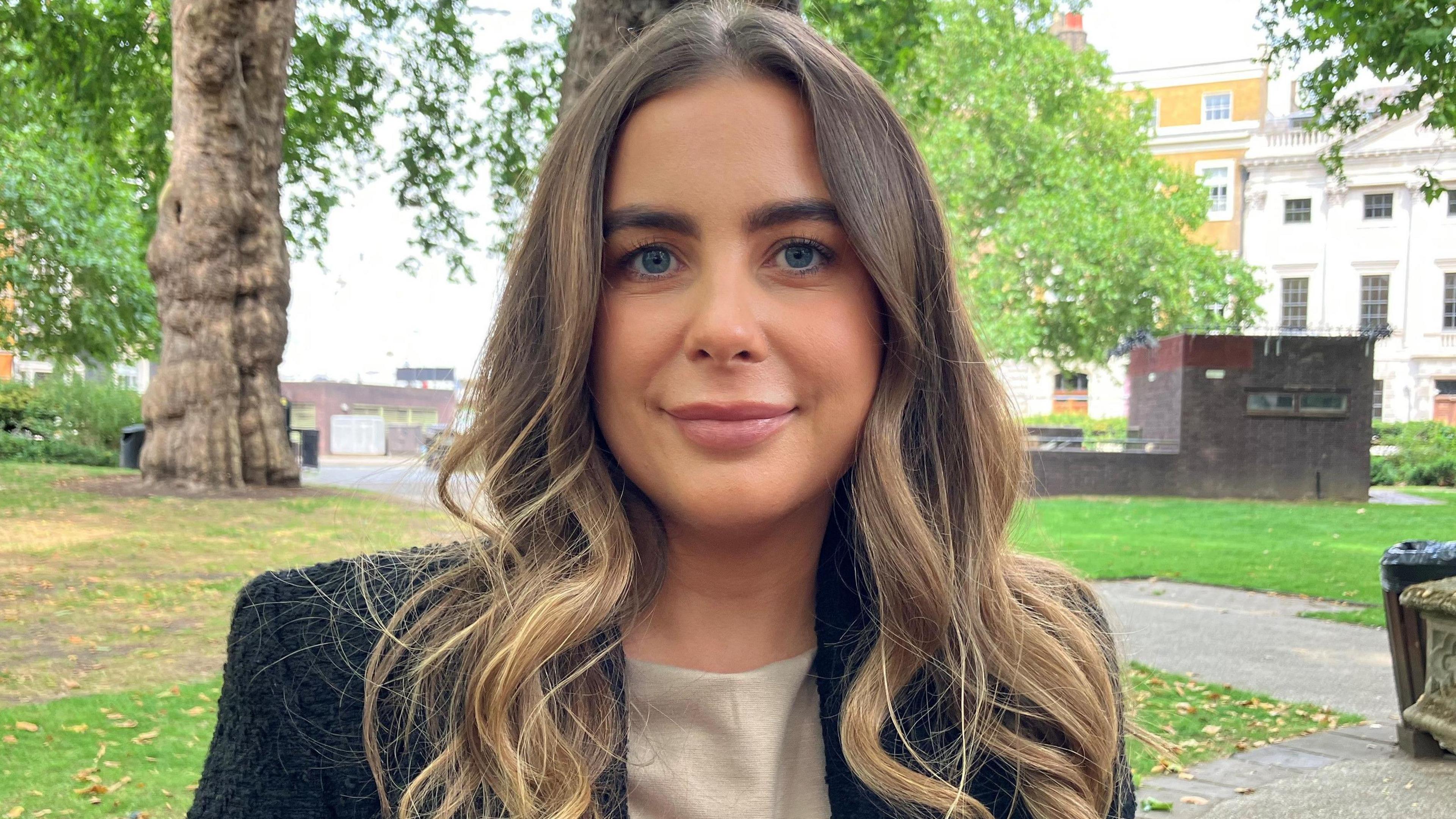 Woman stands in a park with trees and Georgian-style housing behind her. She look at the camera. She has long brown-blond hair and wears a dark suit jacket.