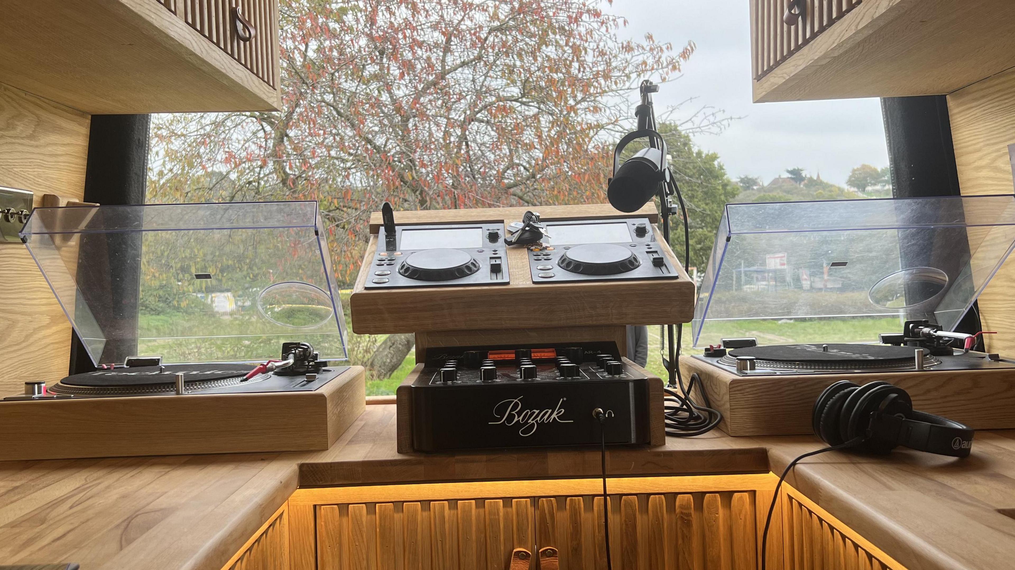 A close up of Mr Lydon's DJ decks, which are sitting on top of a wooden cabinet. There are two record players and a microphone. The van's doors are open with a view of a green playing field and tree. 