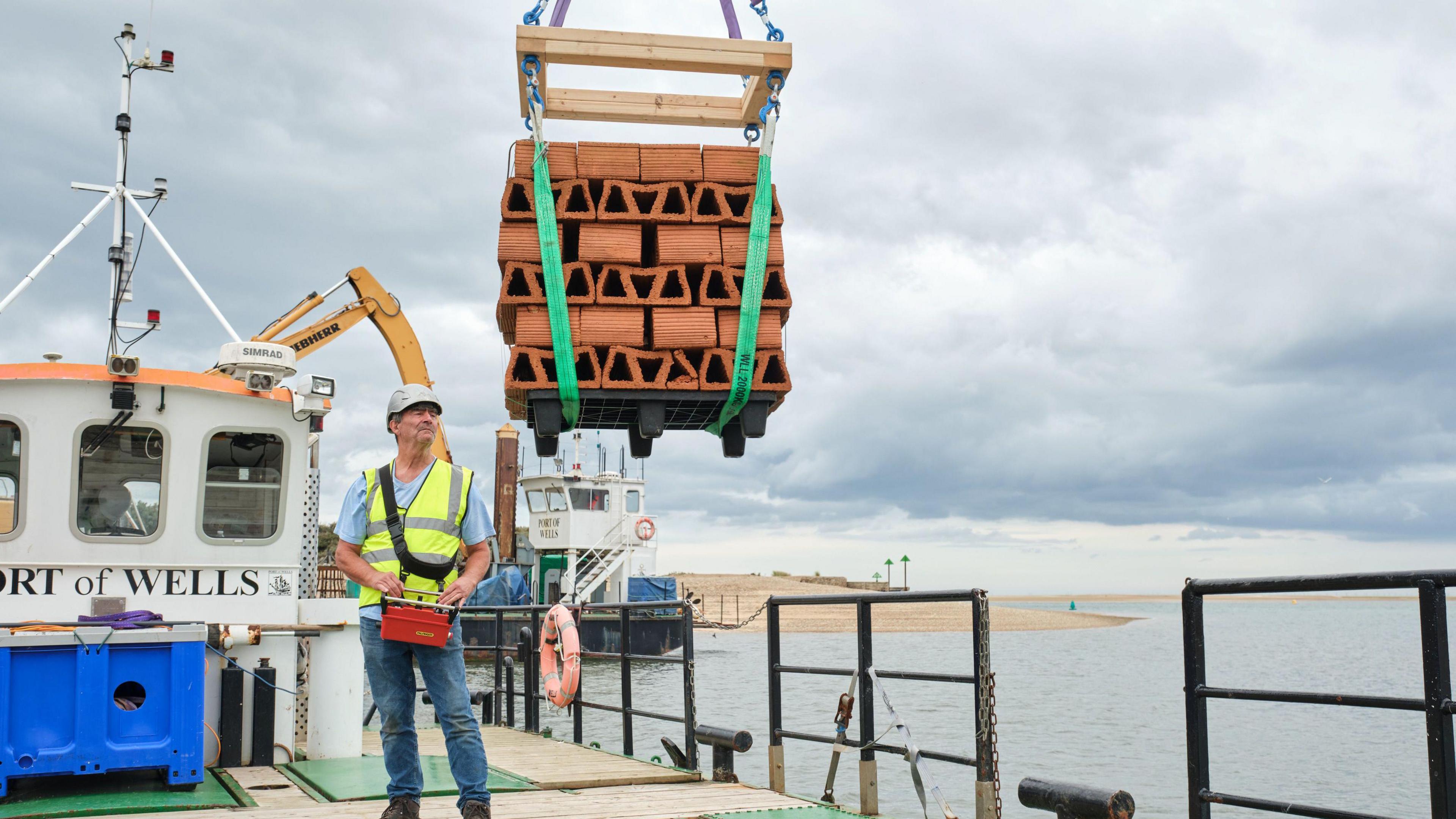 A person uses a crane to lift a pallet full of clay bricks from a boat into the sea