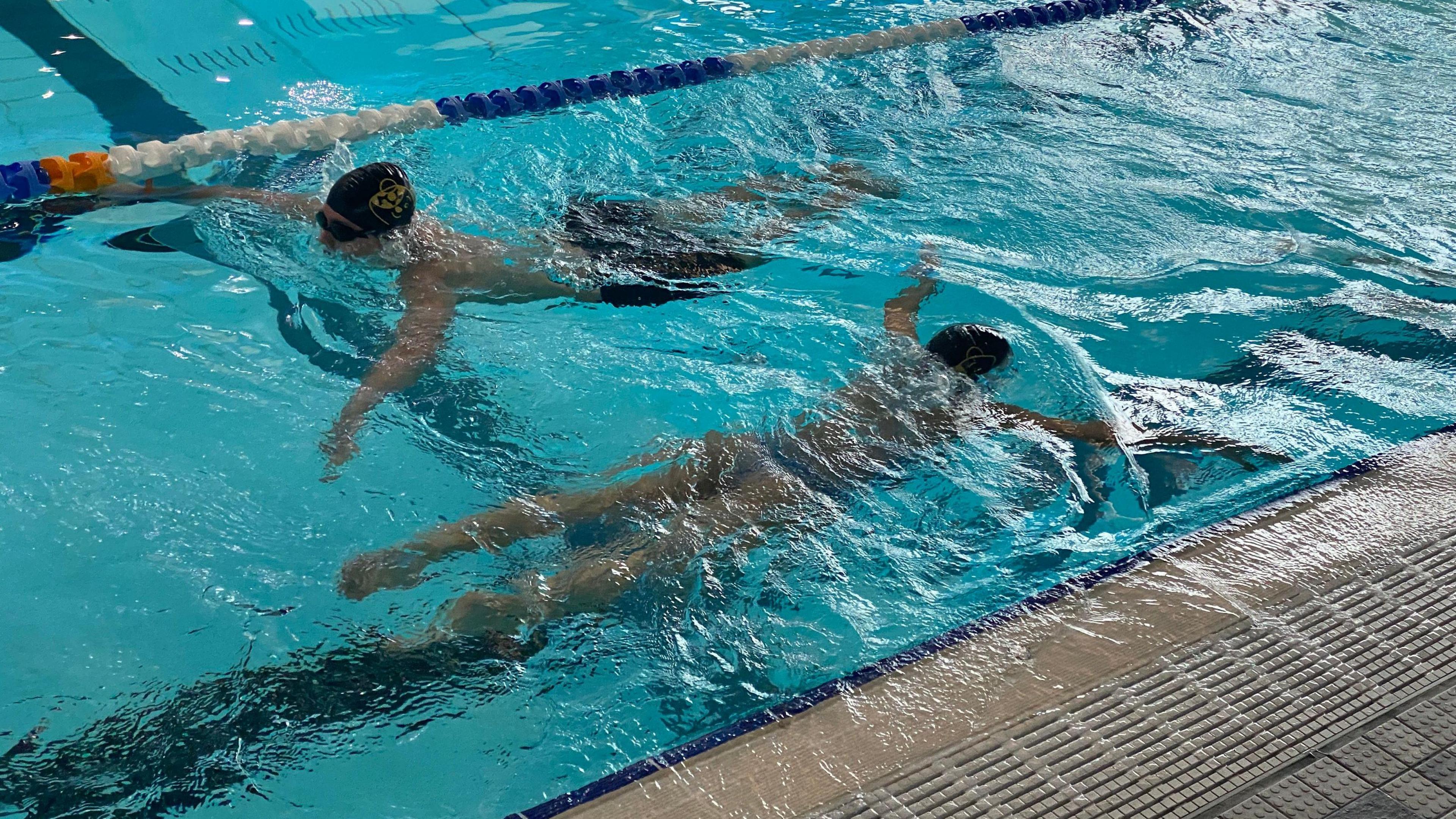 Both Gemma and Luca are in the water in the pool and the camera is over the top of them looking down. They're both doing breast stroke. Luca is wearing black swimming trunks and Gemma is in a light blue and patterned swim costume.