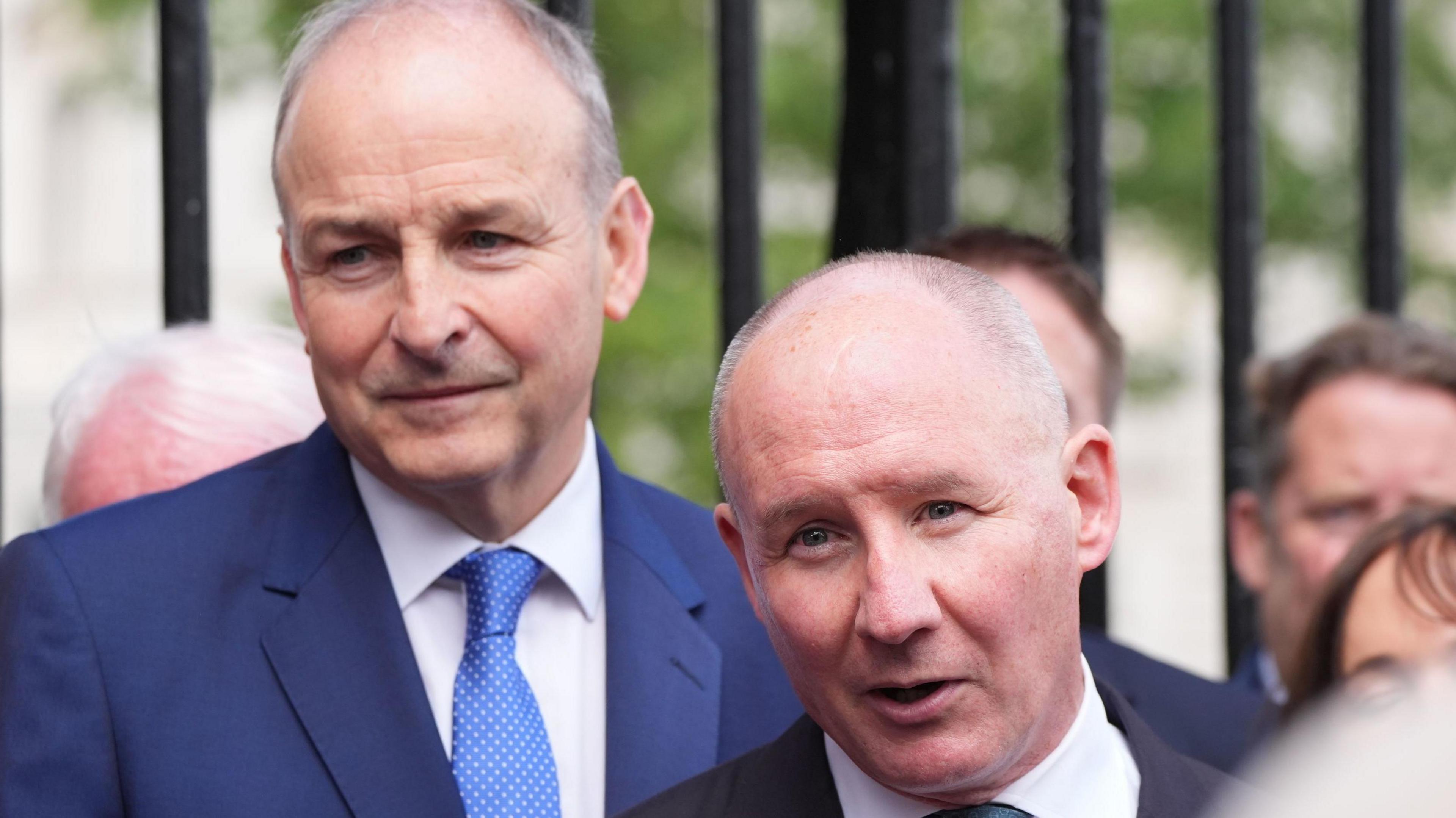 Jim Gavin (right) and Micheal Martin (left), both in blue suits, with white shirts and blue ties. They each have grey hair. There are black railings behind them.