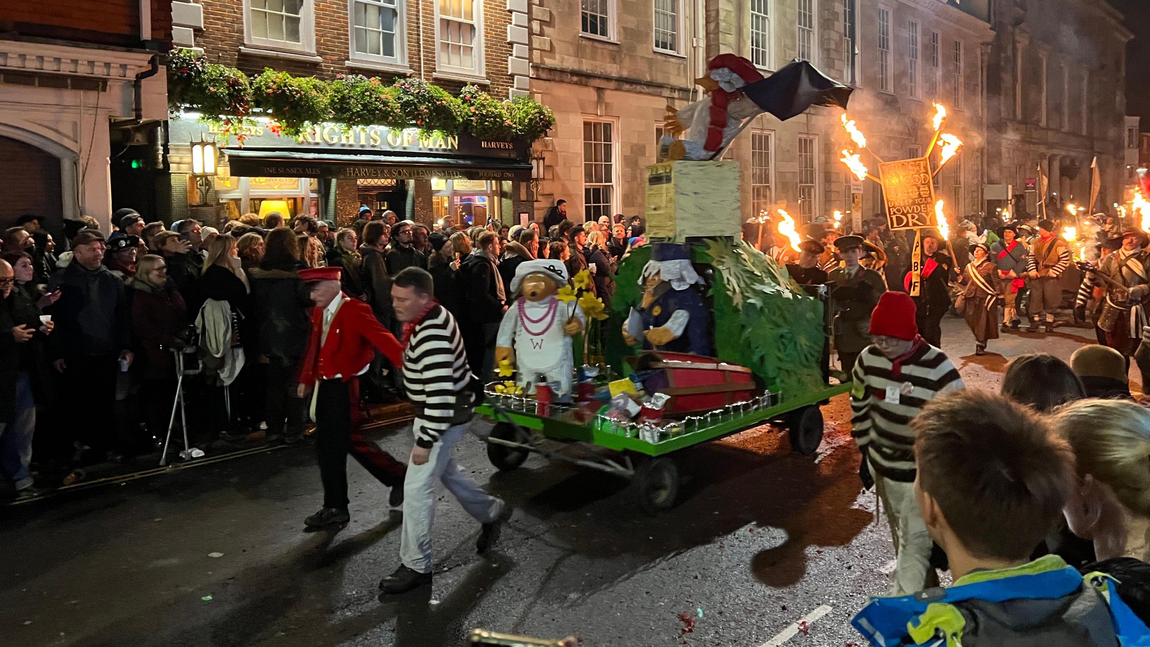 Members of the Cliffe Bonfire Society walking through Lewes High Street with an effigy and torches following behind as crowds watch on.