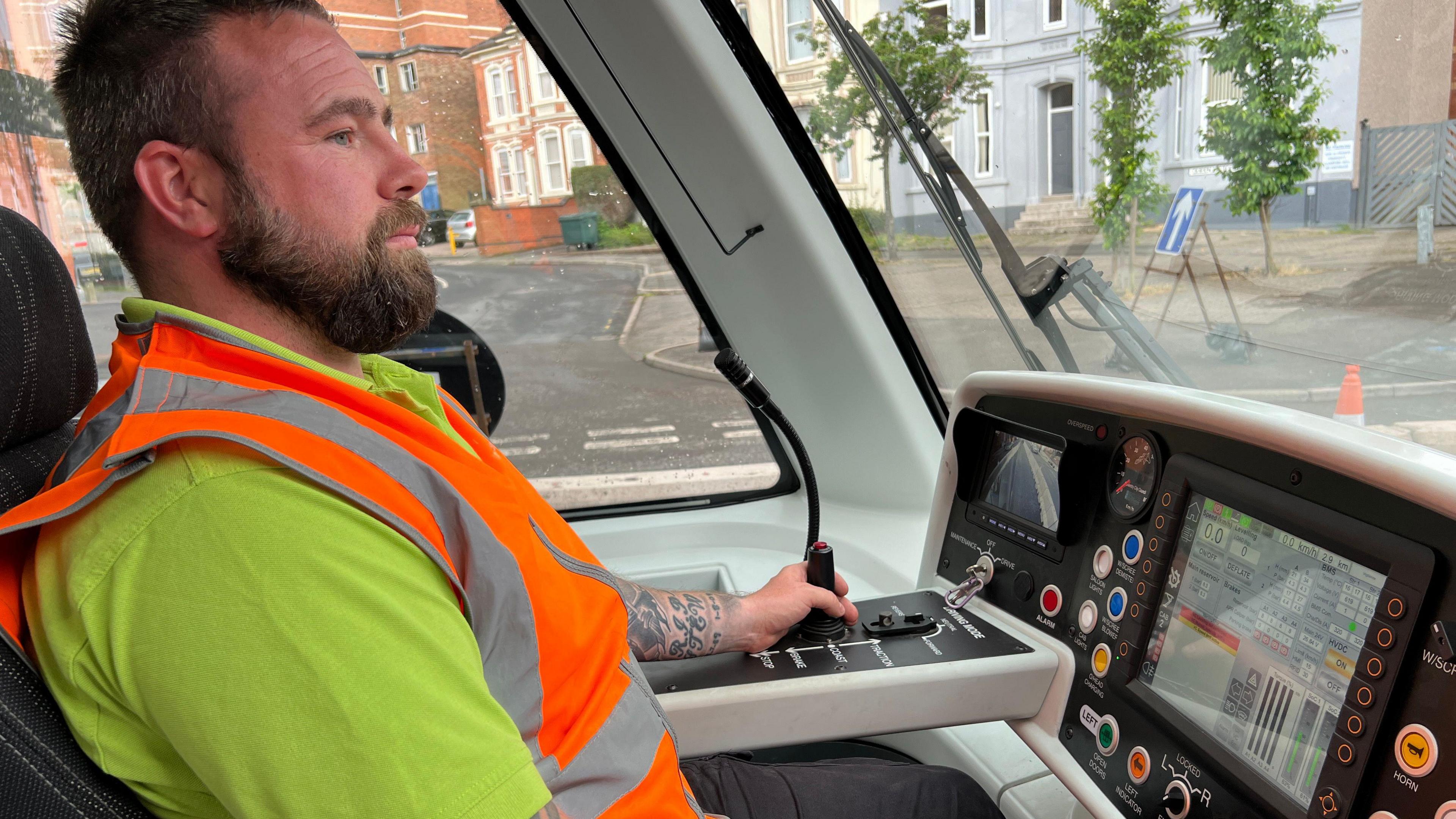 A man in a high-vis vest drives a Very Light Rail vehicle. He has one hand on the dashboard operating the controls and another on his knee. He is looking at the way ahead.