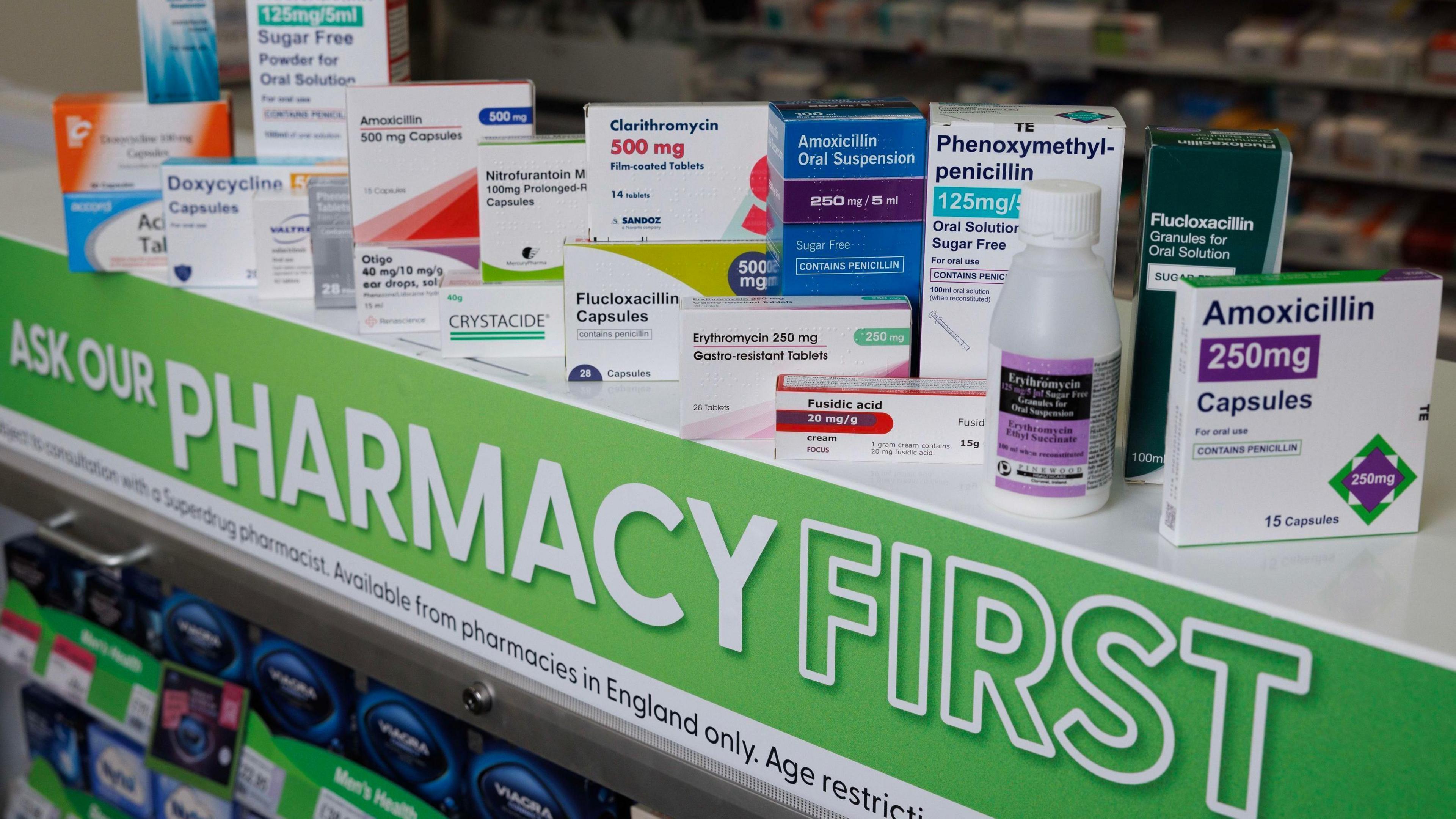 24 different types of medications that Superdrug is now able to supply to patients under the Pharmacy First service. They are all lined up on a shelf with "Ask our Pharmacy First" printed in white letters on a green sign.