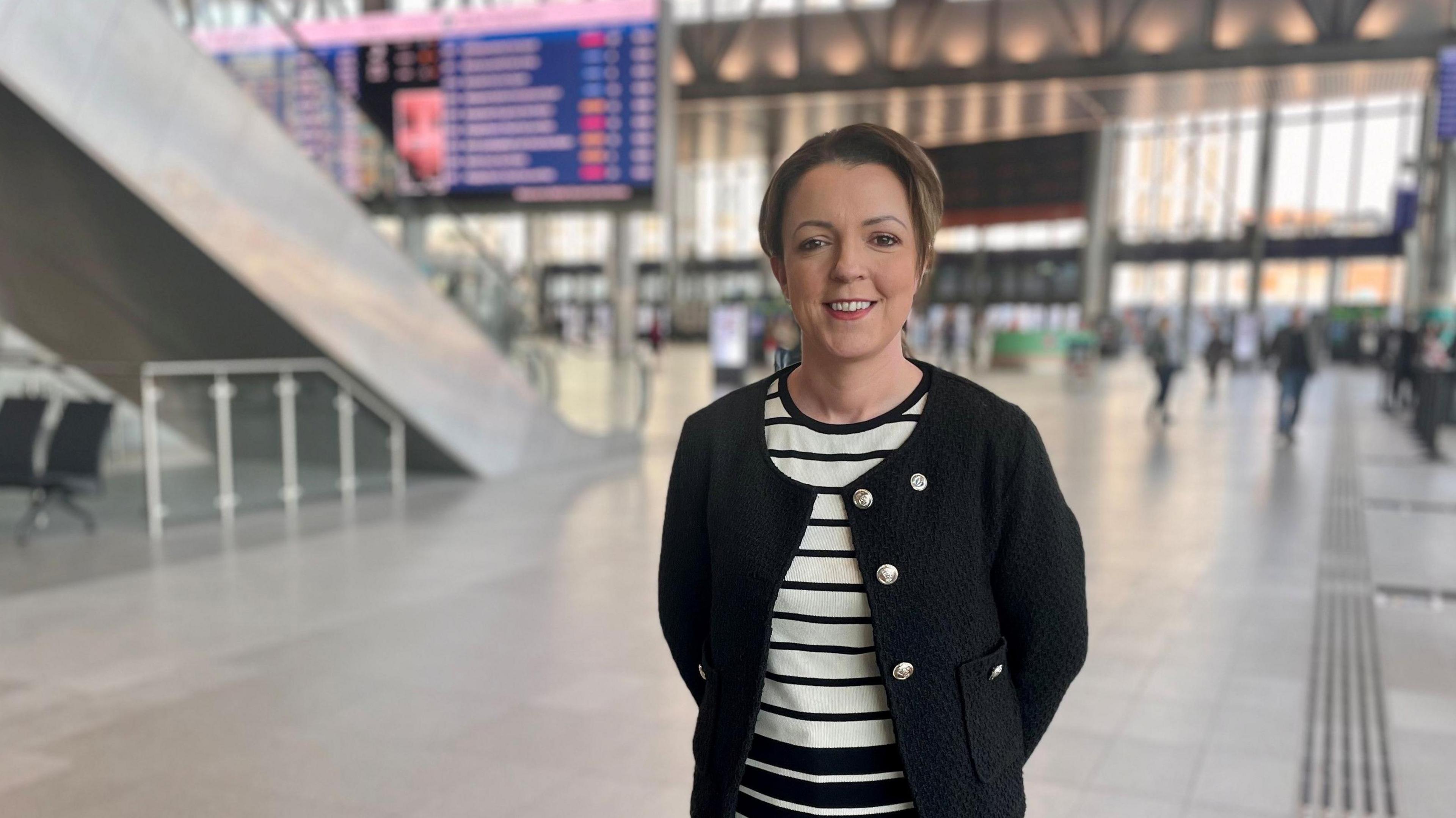 Liz is standing in a station, behind her is a screen and people blurred out. She is wearing a black blazer and white and black t-shirt underneath. She has blond hair tied back in a ponytail. She is smiling at the camera with her hands behind her back. 