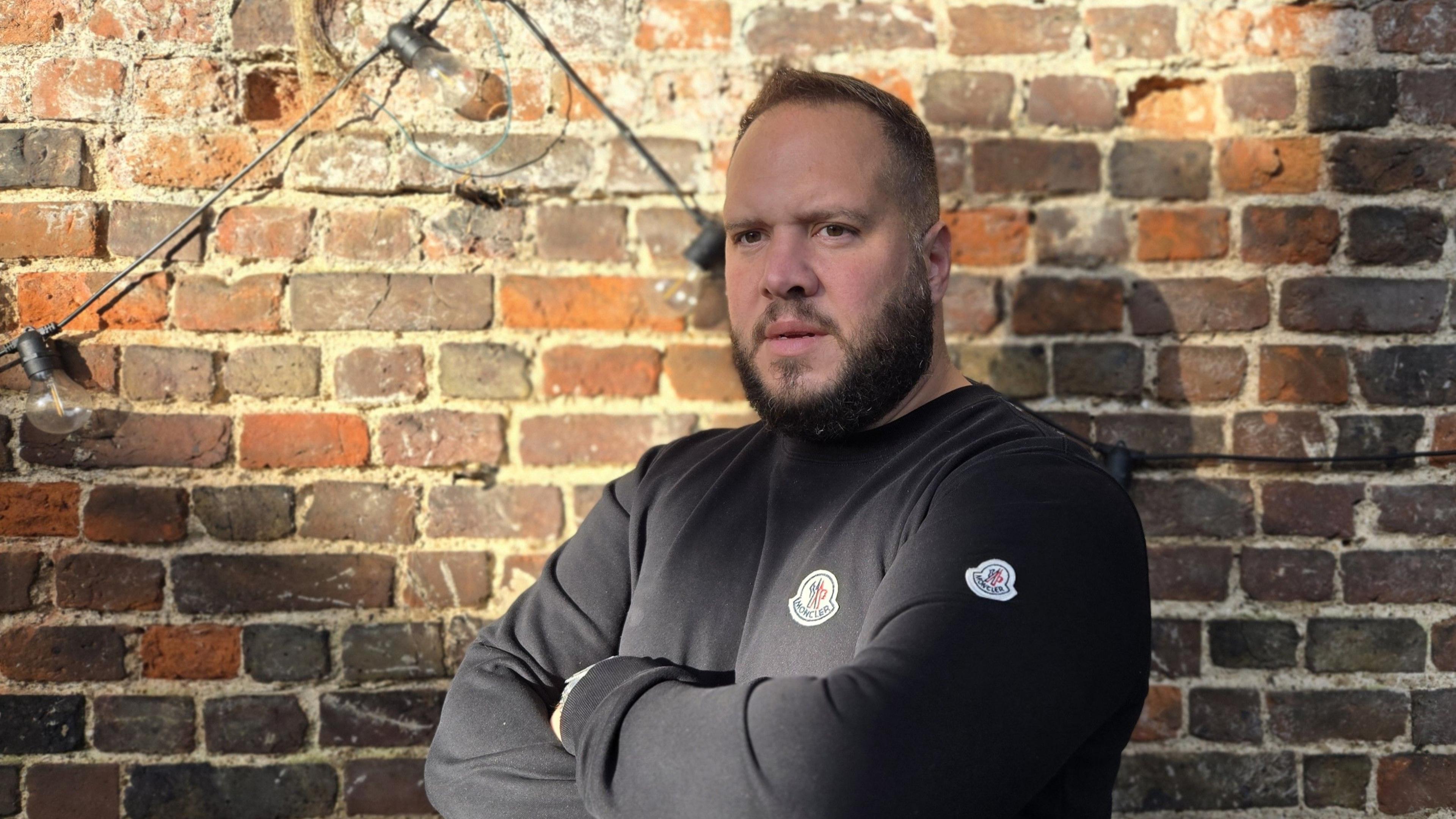 Chris Camacho in a black, long-sleeved, collarless shirt, stands with his arm folded in front of an old brick wall.