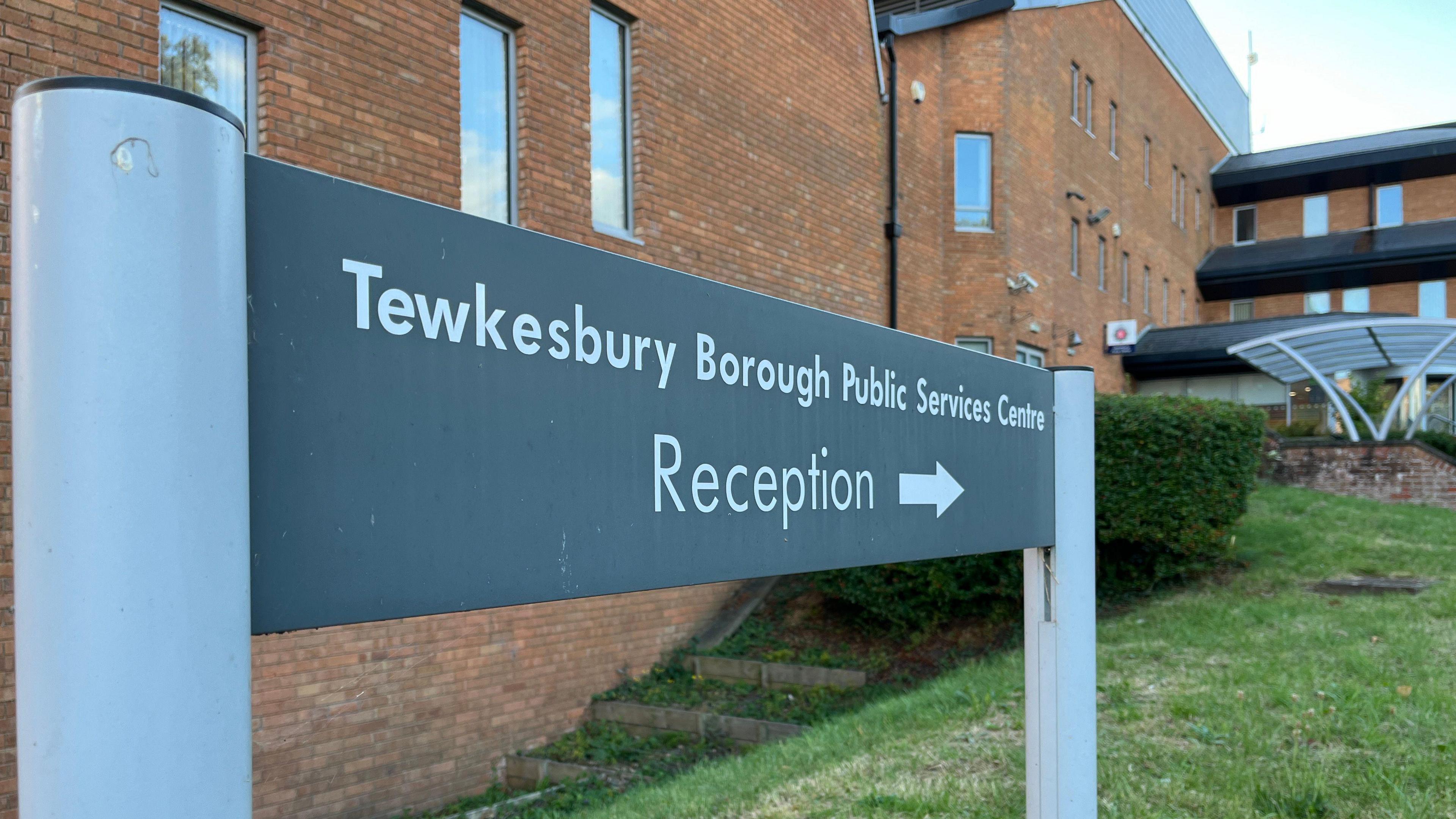 A grey sign in front of the entrance to Tewkesbury Borough Council which points to the reception. There is a red brick building behind and grass and a bush in the foreground. 