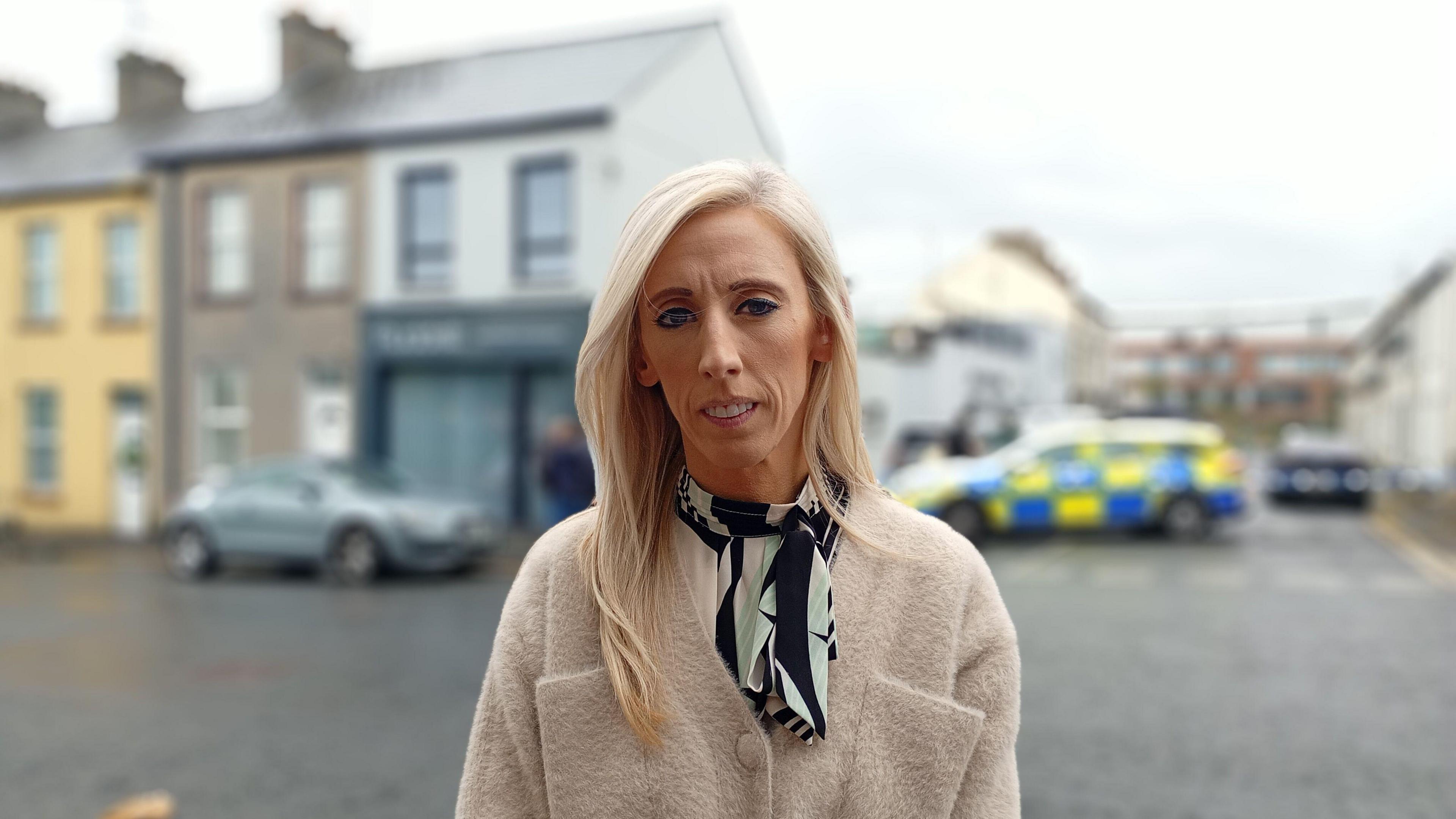 Carla Lockhart, a woman with long, straight, blonde hair, looks directly at the camera with a serious expression while standing close to the scene of the car fire in Portadown.   She is wearing a beige cardigan with large front pockets and black and pale green scarf tied around her neck.  The background is out of focus but there are houses and a parked police car behind her. 