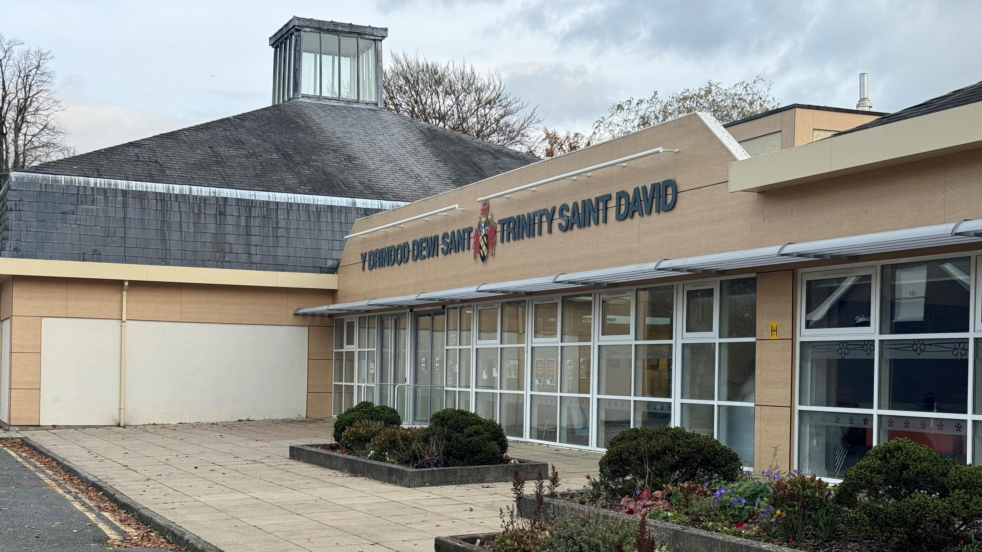 A light building with large glass windows from the floor with a grey roof, with a sign reading Y Drindod Dewi Sant Trinity Saint David