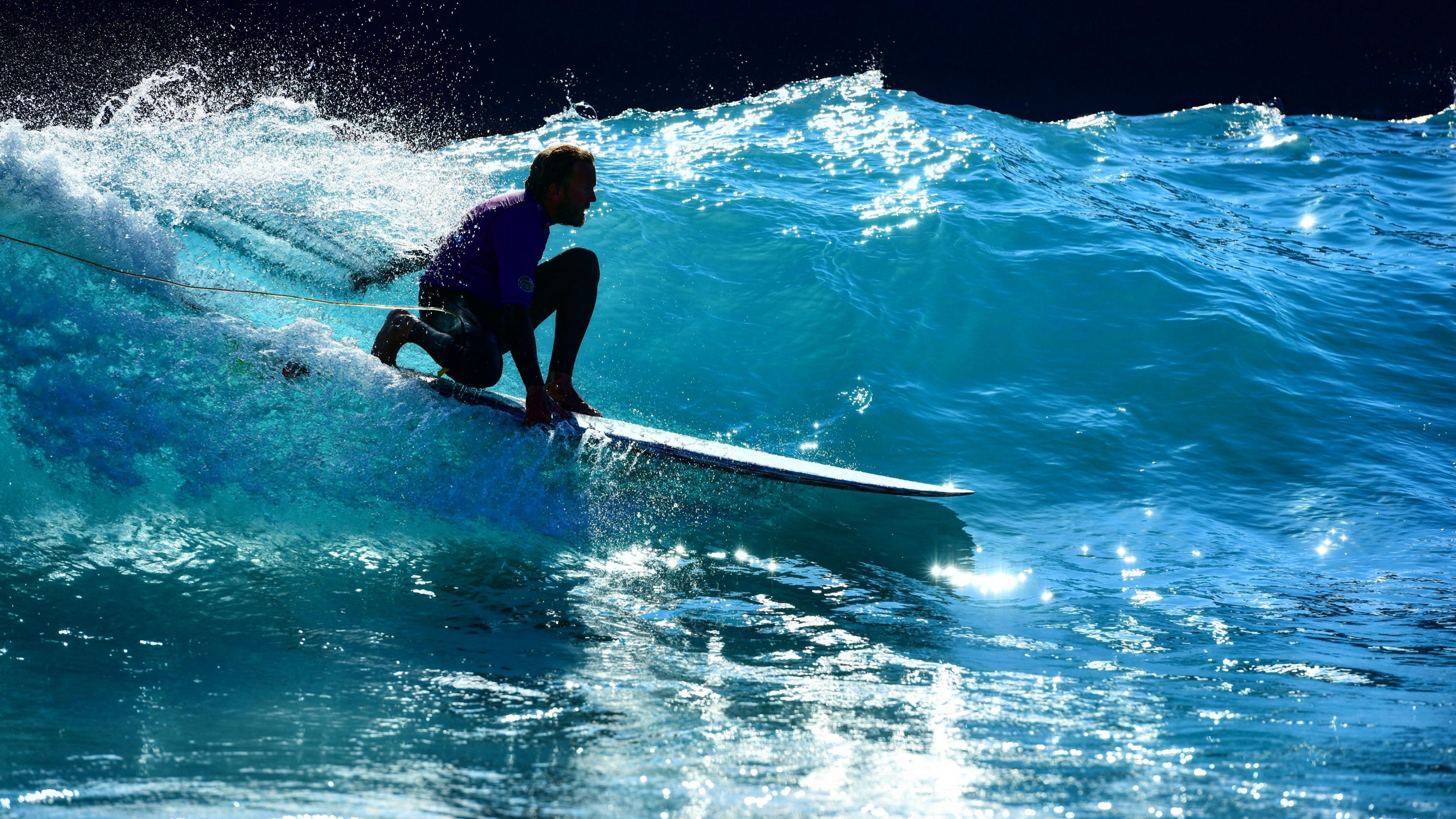 Nick Hounsfield surfing along an unbroken wave on a white longboard. He is wearing a black wetsuit and purple rash vest, and is crouched down low on the board. The bright blue water is sparkling in the sunlight. 