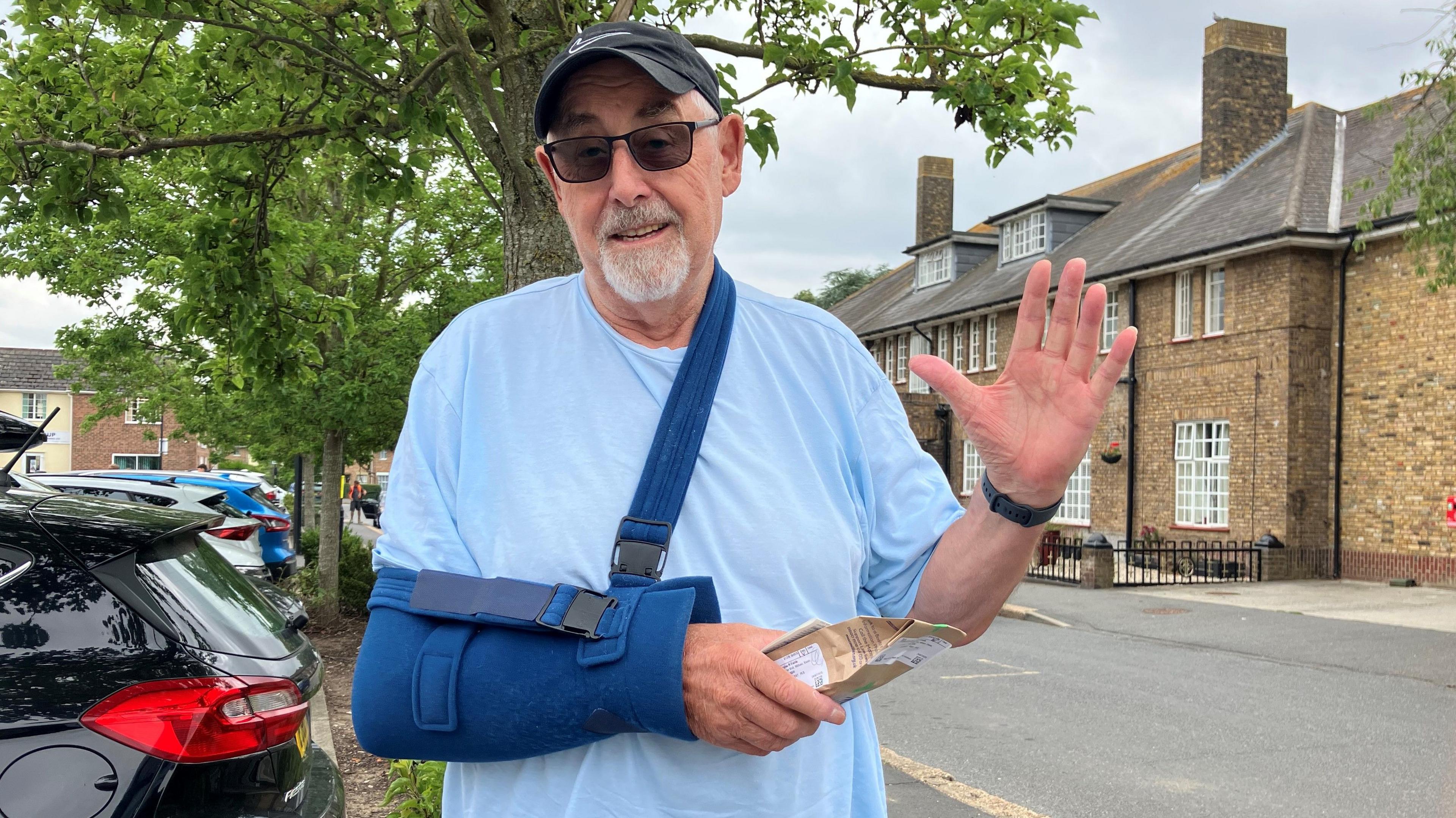 Doug Fields in a blue T-shirt, sunglasses and wearing a dark blue cast on one arm, while he looks to be waving or gesturing at the camera with the other arm.