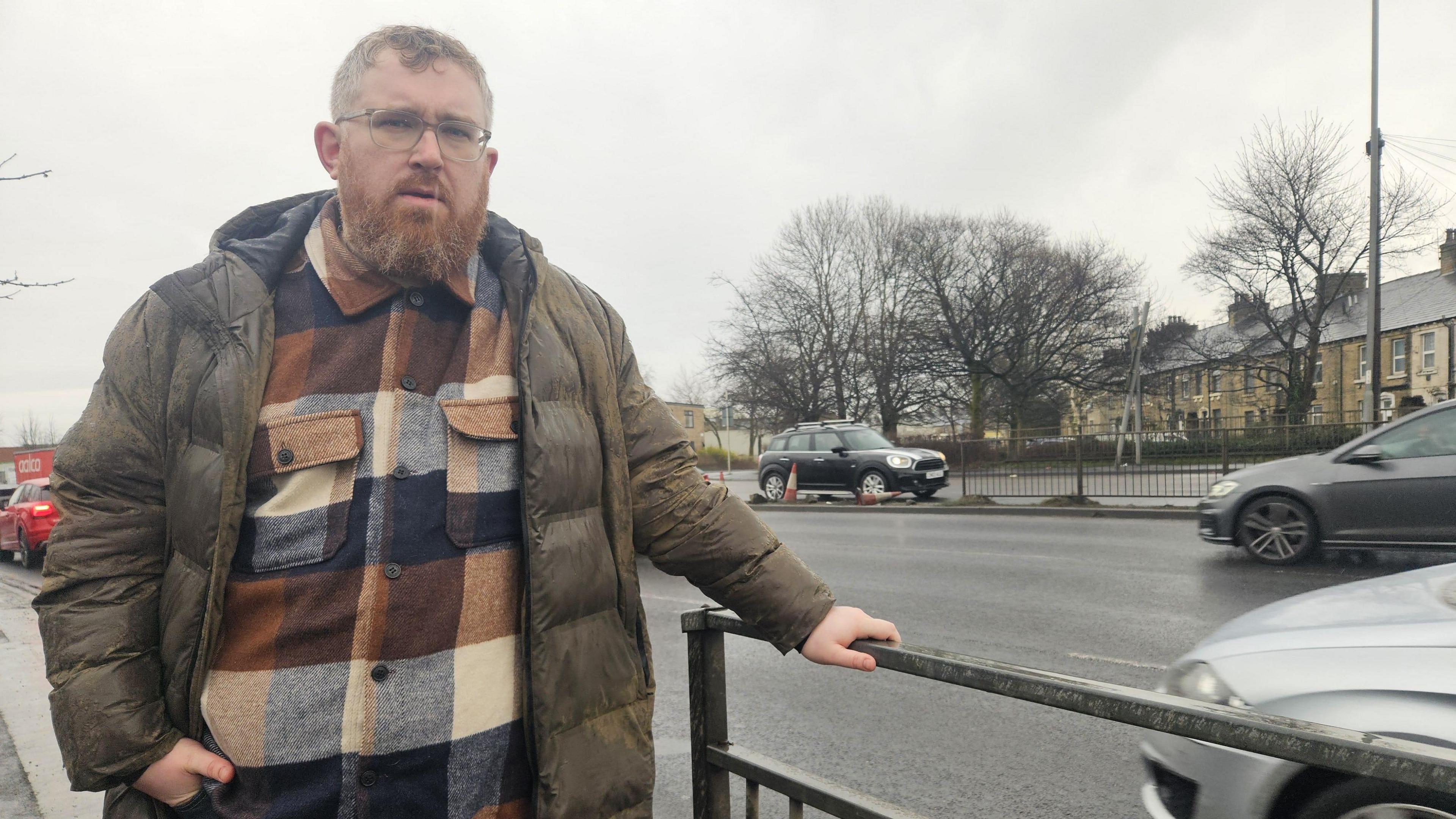 A man stands in front of some metal barriers on a dual carriageway road. Behind him, traffic rushes past and a section of the middle crash barrier is  missing, with traffic cones marking it off.