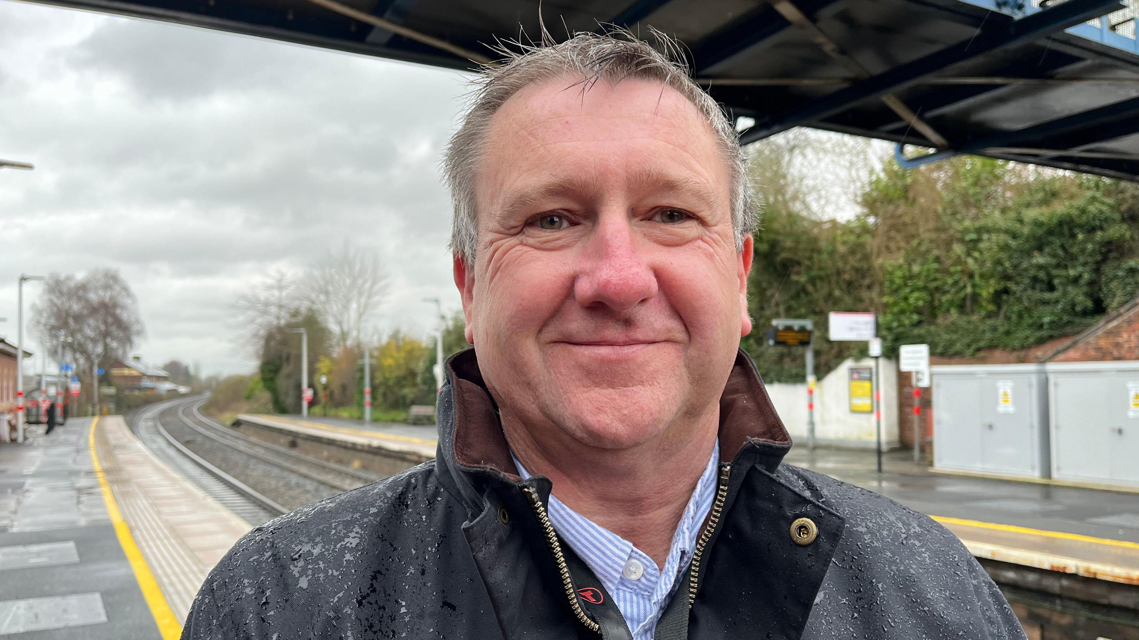 Adrian looking into the camera and smiling. He's wearing a black waterproof coat - with raindrops on the shoulders as it's clearly bad weather - and a blue and white striped shirt underneath. He's stood on one of Ludlow station's platforms, with the track stretching away into the distance behind him. The sky is grey and overcast.