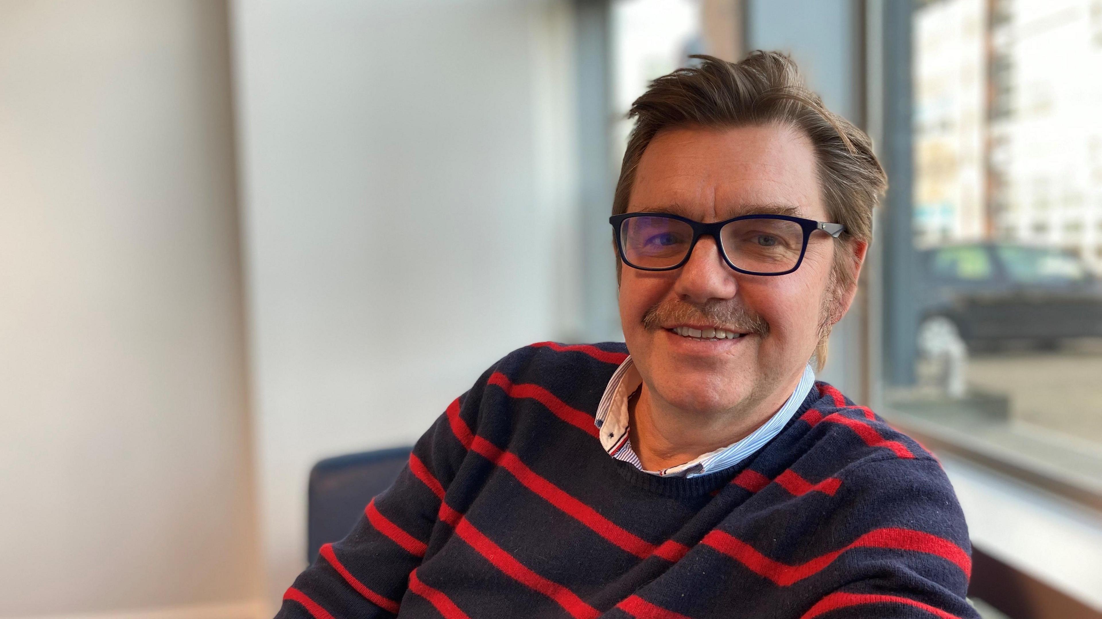 Nik Johnson is wearing a blue and red striped jumper and a shirt. He is looking at the camera smiling, while seated. He has brown hair, a moustache and thick framed glasses.