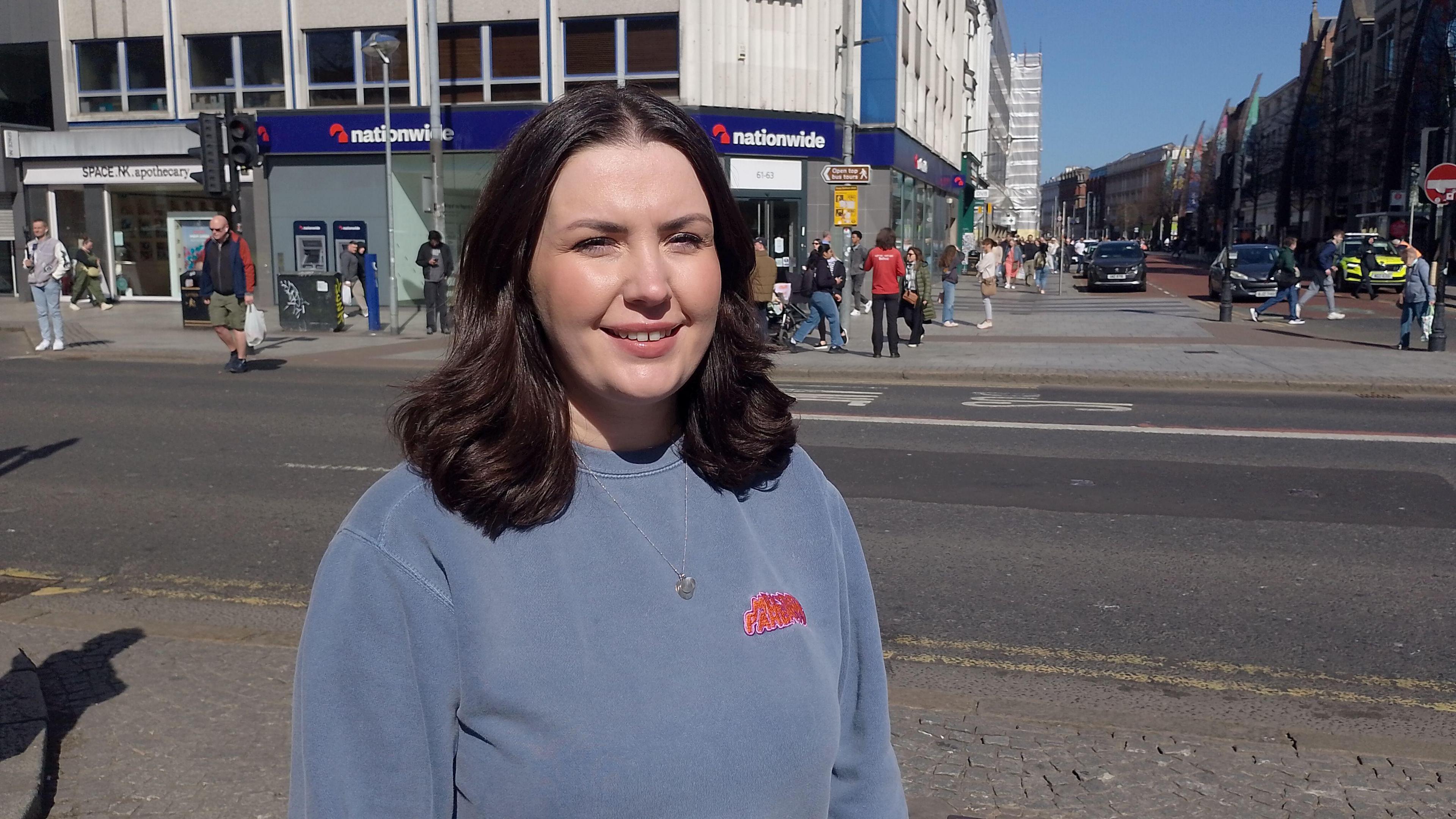 A woman, with shoulder length brown hair, looking at the camera. She is wearing a blue jumper. She's standing on a path with a road and a Nationwide bank in the background.
