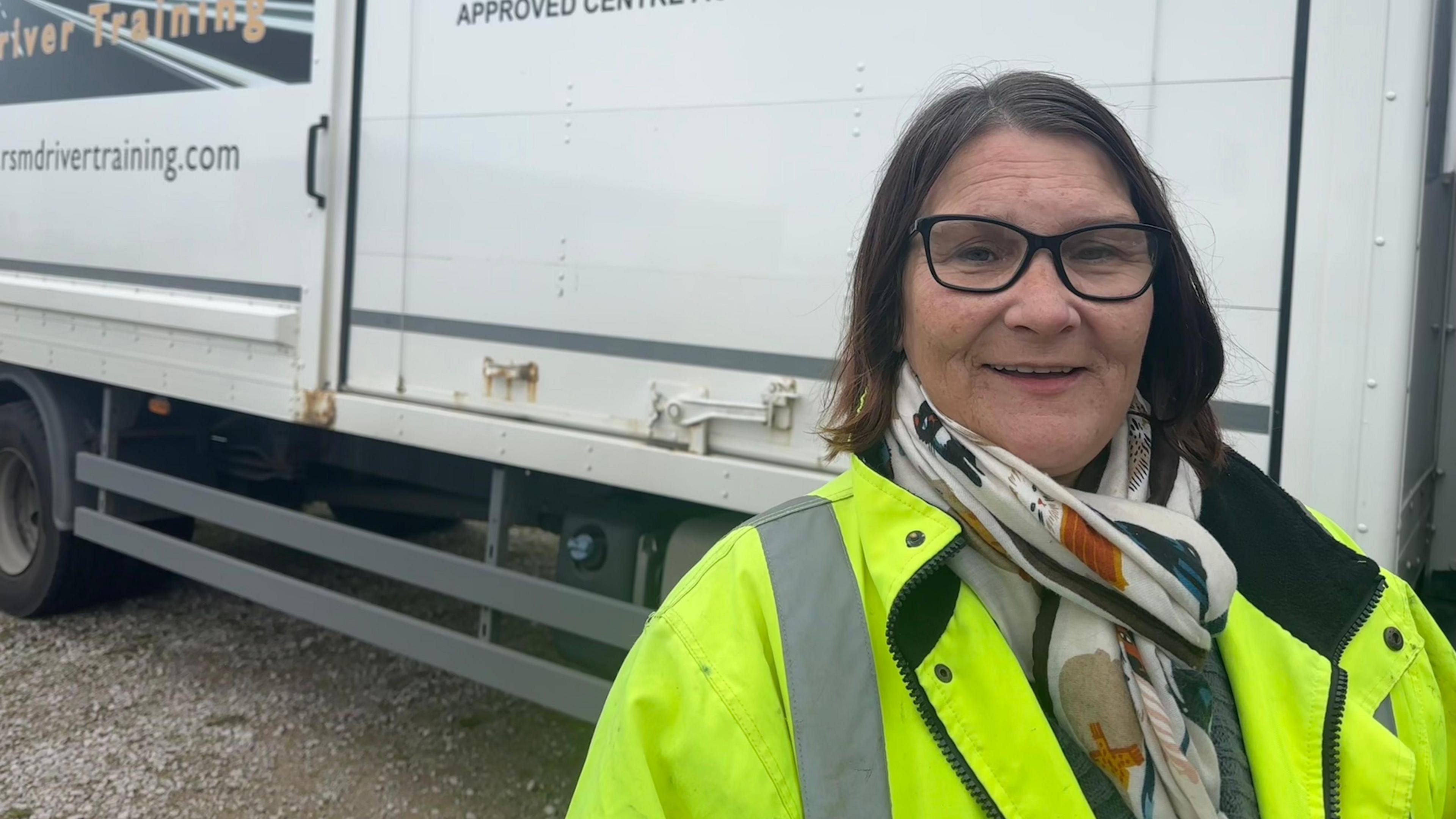 Theresa with brown hair wearing glasses, a scarf and a hi-vis jacket smiling into the camera