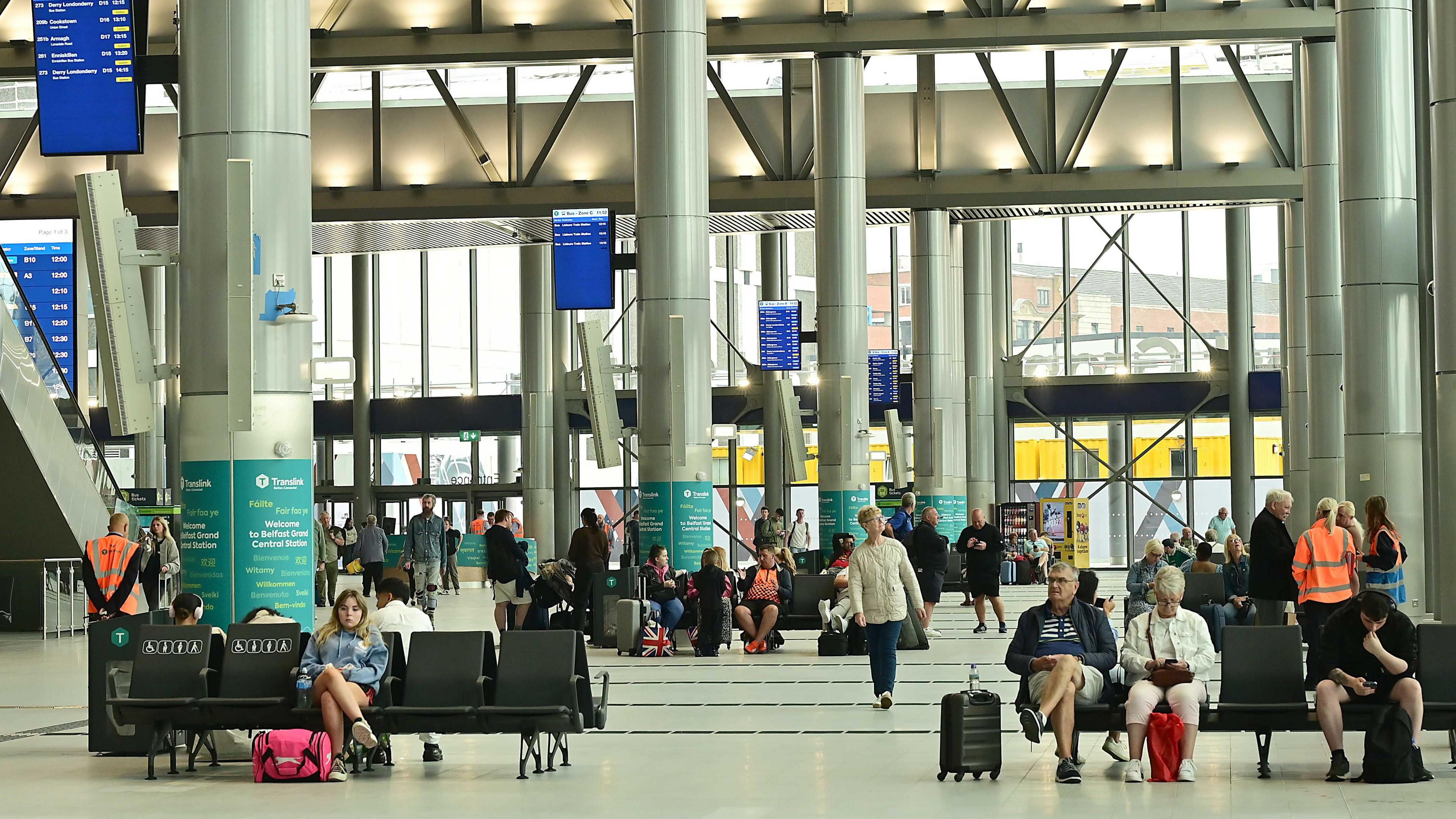 The foyer area of the new station. Passengers are sitting in seating areas waiting for their buses. Some have suitcases and travel bags.