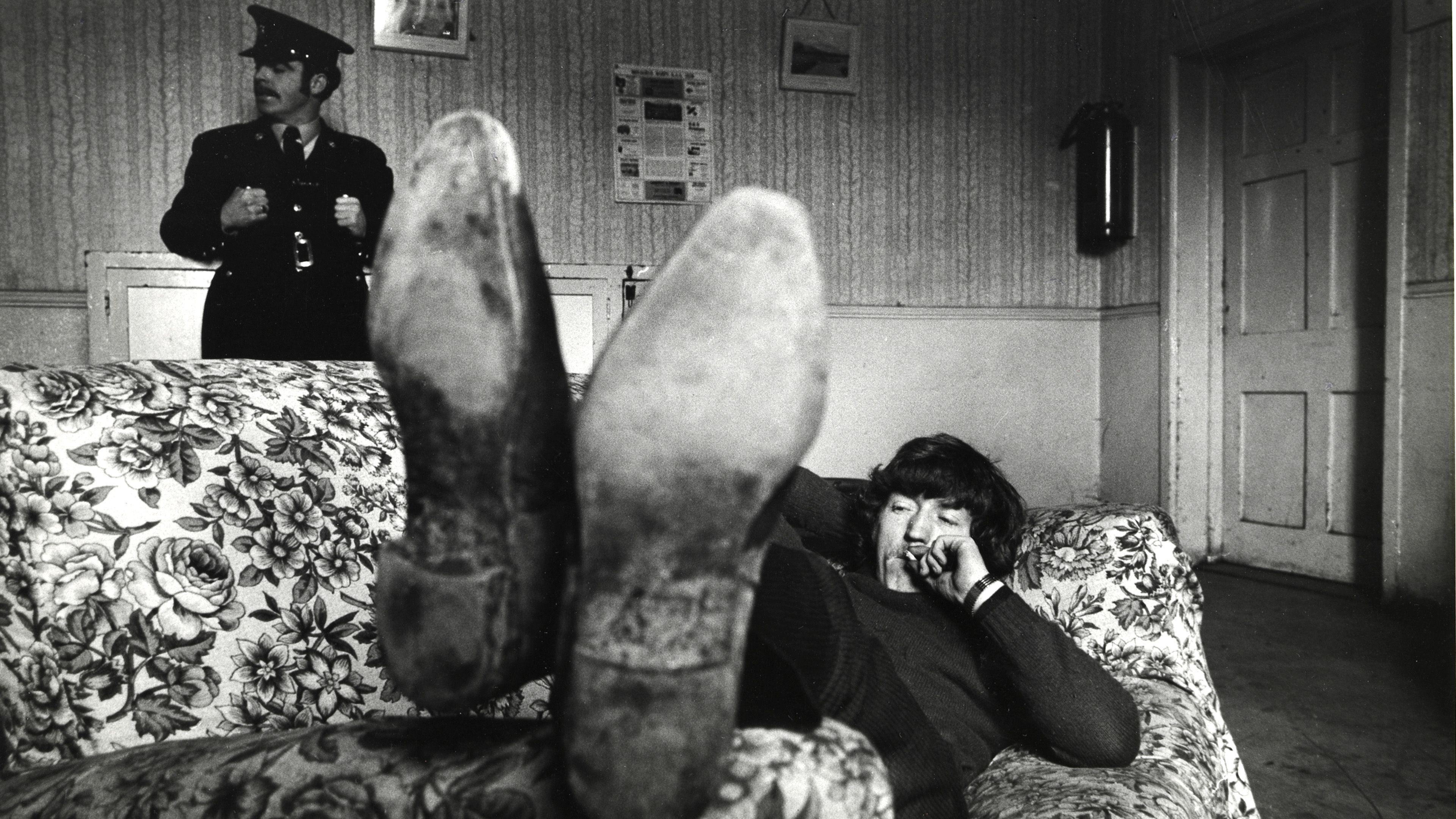 Brendan is pictured laying on a floral couch as a young man. The bottom of his scuffed up formal shoes are close to the camera on the armrest. Brendan lays back in dark jeans and a jumper with one arm behind his head. He is smoking a cigarette with the other hand, with a watch poking out of his sleeve. He has medium length dark hair which is swooped to the side of his face. In the background of the image a scuffed up white door is to Brendan's right, while a police officer stands to his left. He is behind the couch but in front of a wood panel wall. The officer is dressed in an old style black police uniform. It has large metal buttons and a belt buckle on show. 