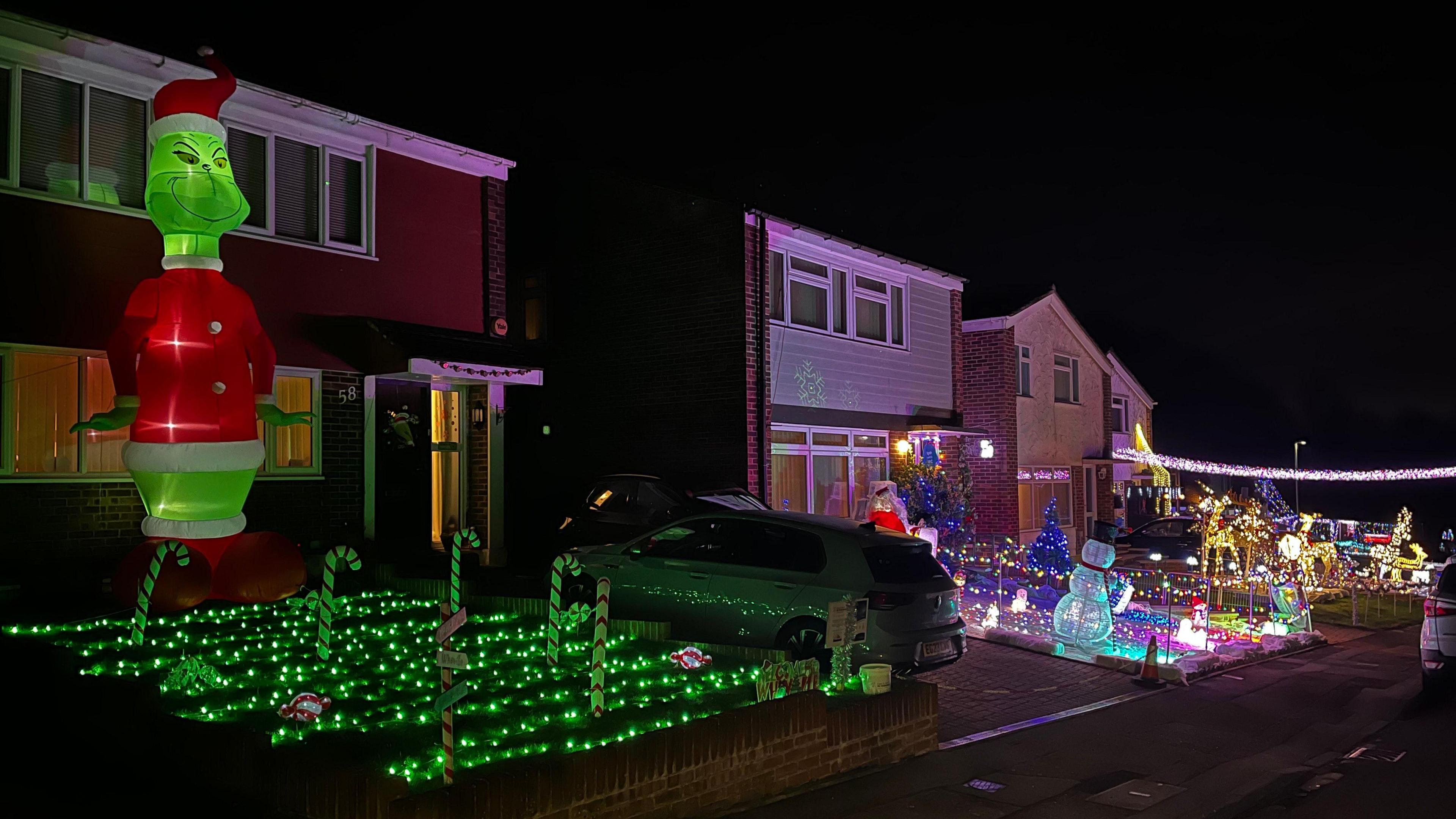 An inflatable Grinch, which stands as tall as a two-storey house, stands outside a property where the front garden is filled with green fairy lights and half a dozen candy canes. Further down the road there are other properties whose gardens are filled with lights and decorative figurines.