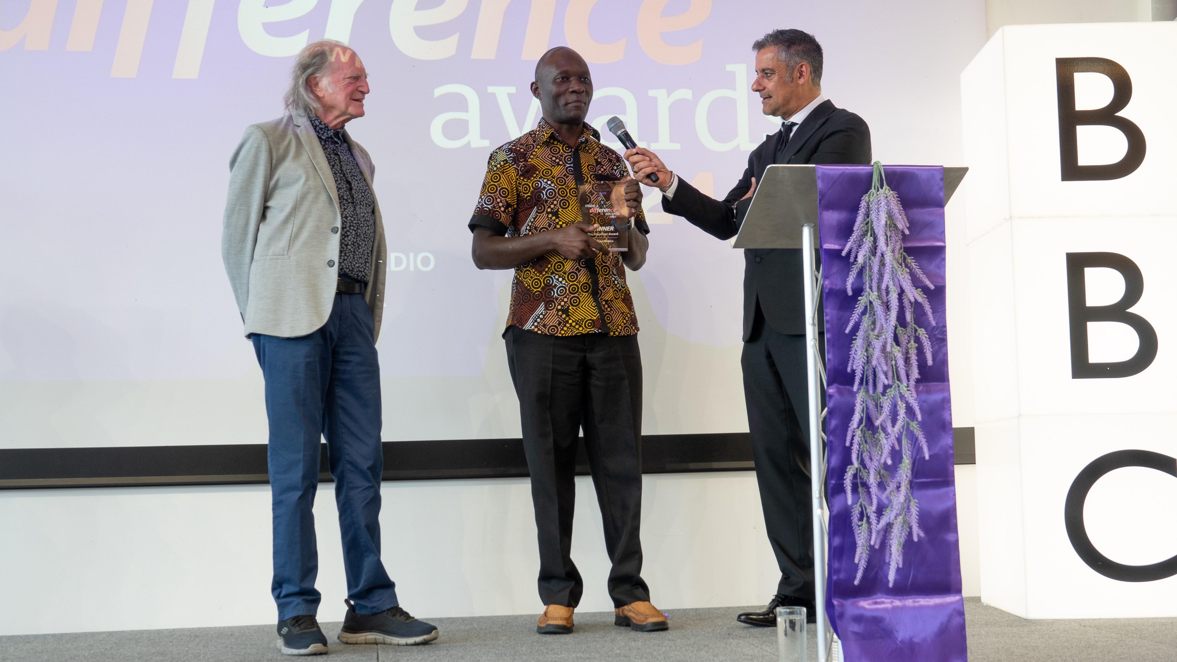 A man receives an award on stage, in front of a screen. There is a large light-up sign that reads BBC. There is a purple podium. A man is holding a microphone to the award recipient, and another man stands and watches