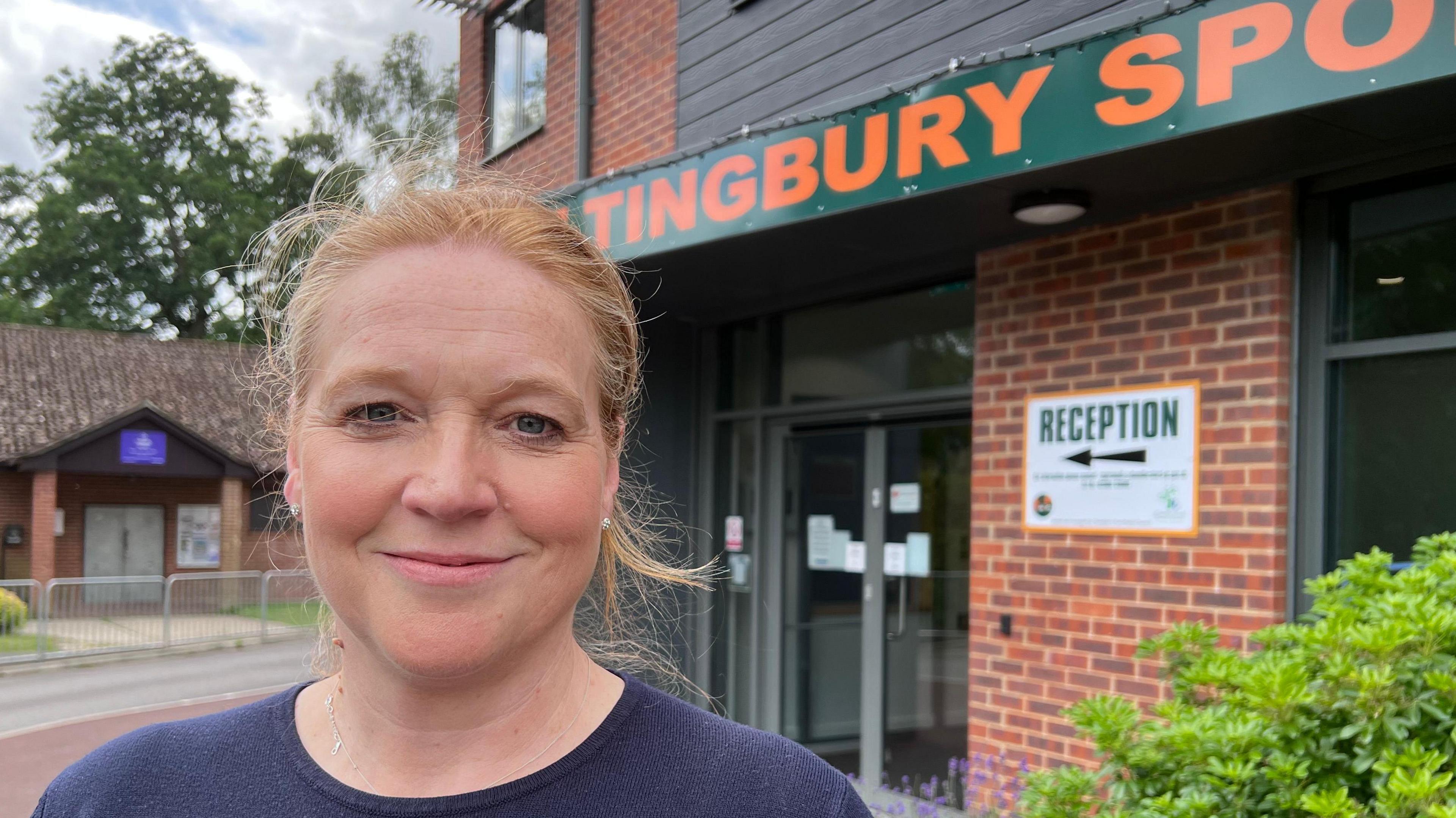 Lady with tied black red hair and a blue jumper stands in front of a two storey brick building. The sign says reception and you can read part of the Hiltingbury sports sign.