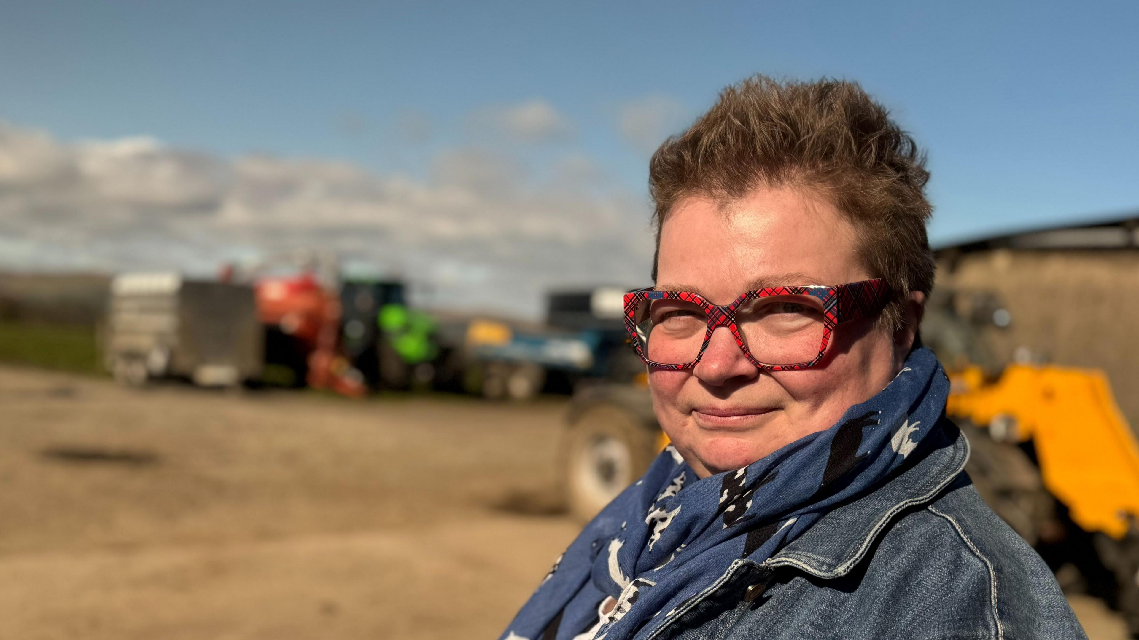 Jennifer Dunlop has short brown hair and is wearing red glasses with a blue scarf and denim shirt. She is smiling at the camera as she stands side on. The background behind her is blurred but there are a range of farming equipment which is red, blue, green and orange coloured.