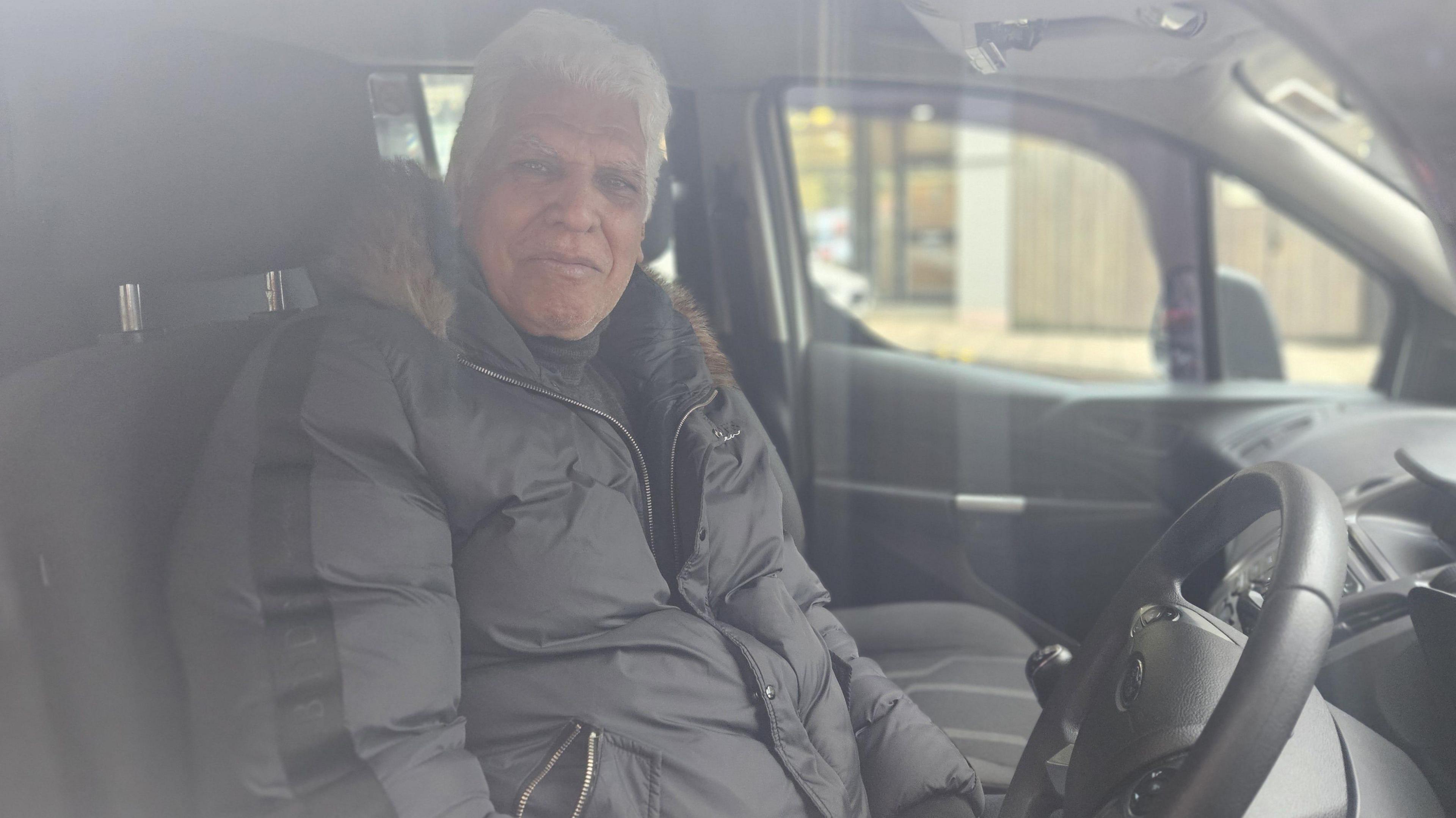 A grey-haired man of South Asian sits in the driver's seat of a car. He wears a puffy black jacket with a fur lined hood.