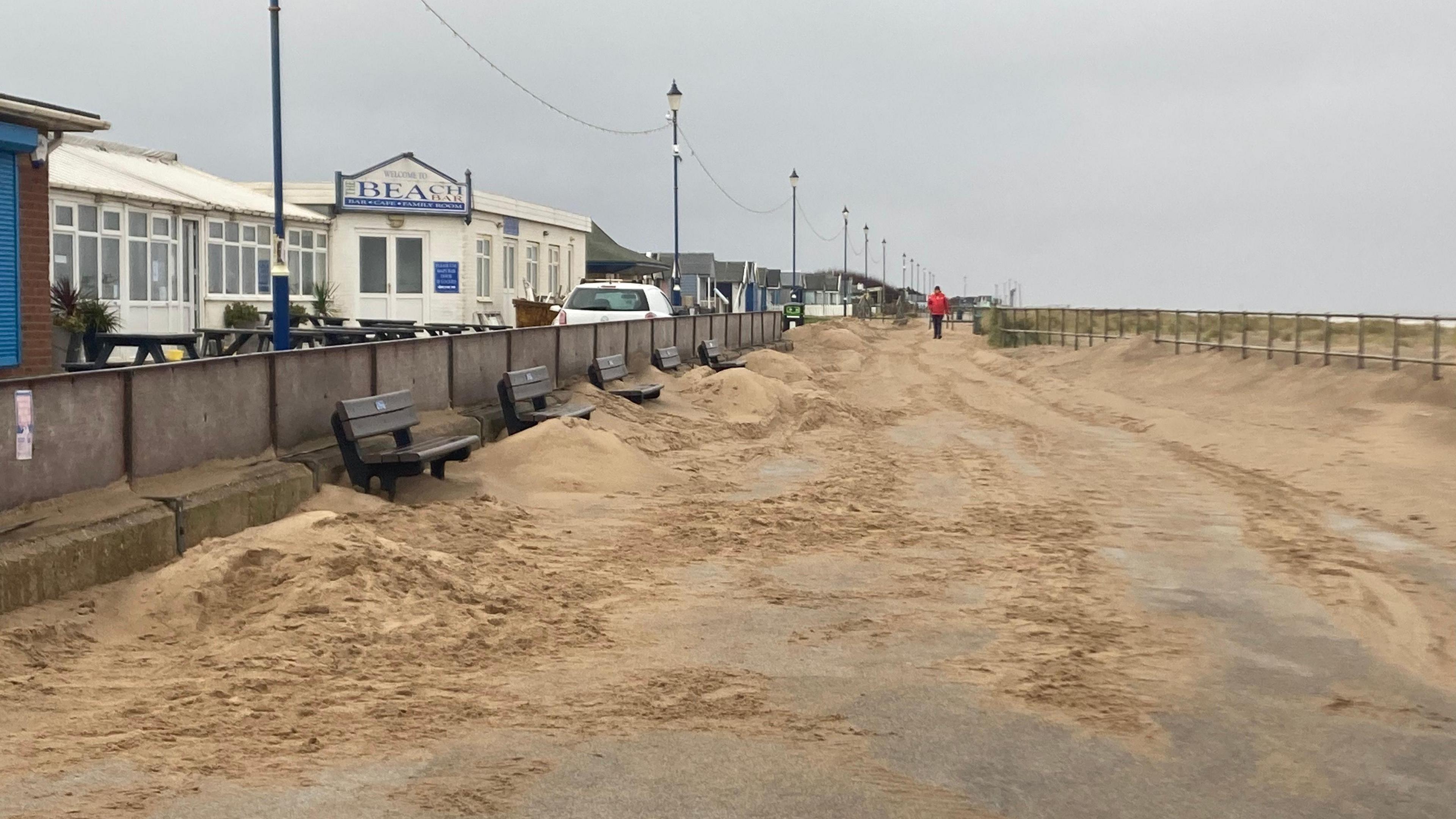 The photo shows a beach promenade. It is covered with piles of sand on either side. There are five benches that are also covered. Along the left hand side, behind a wall, are a row of shops. On the right hand side are some railings which lead onto the sea.