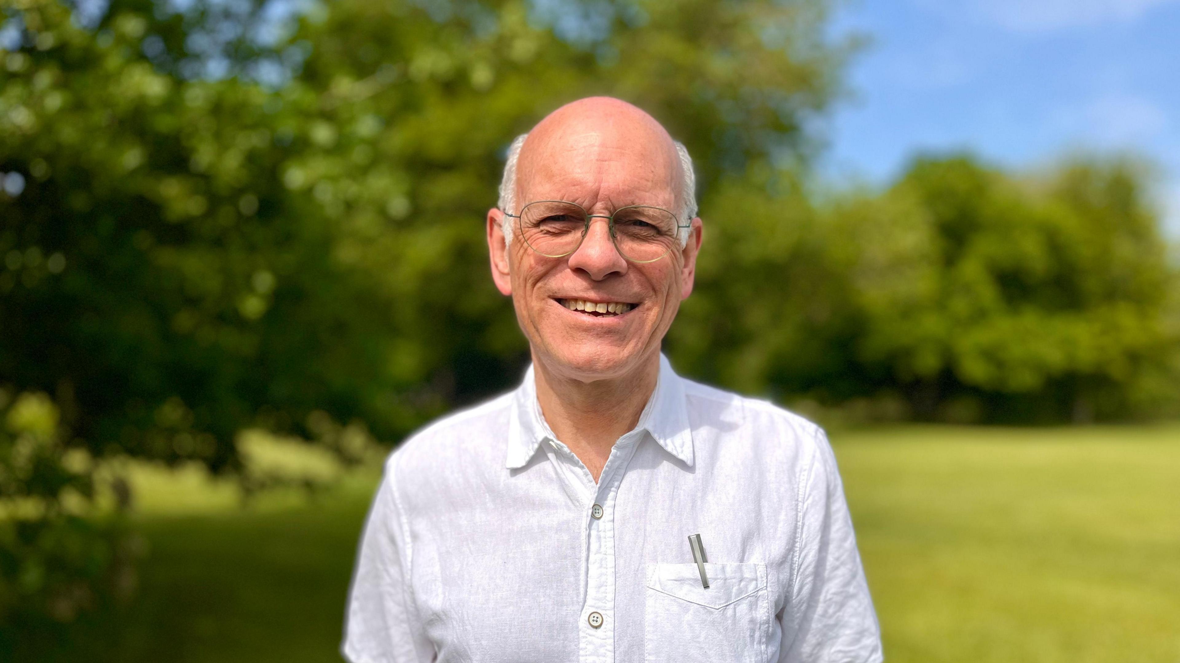 Tim Bick is smiling at the camera. He has white hair around the back and sides of his head and is bald on top. He is wearing glasses and a white shirt, with a pen tucked in his top pocket. In the background is grass and trees.