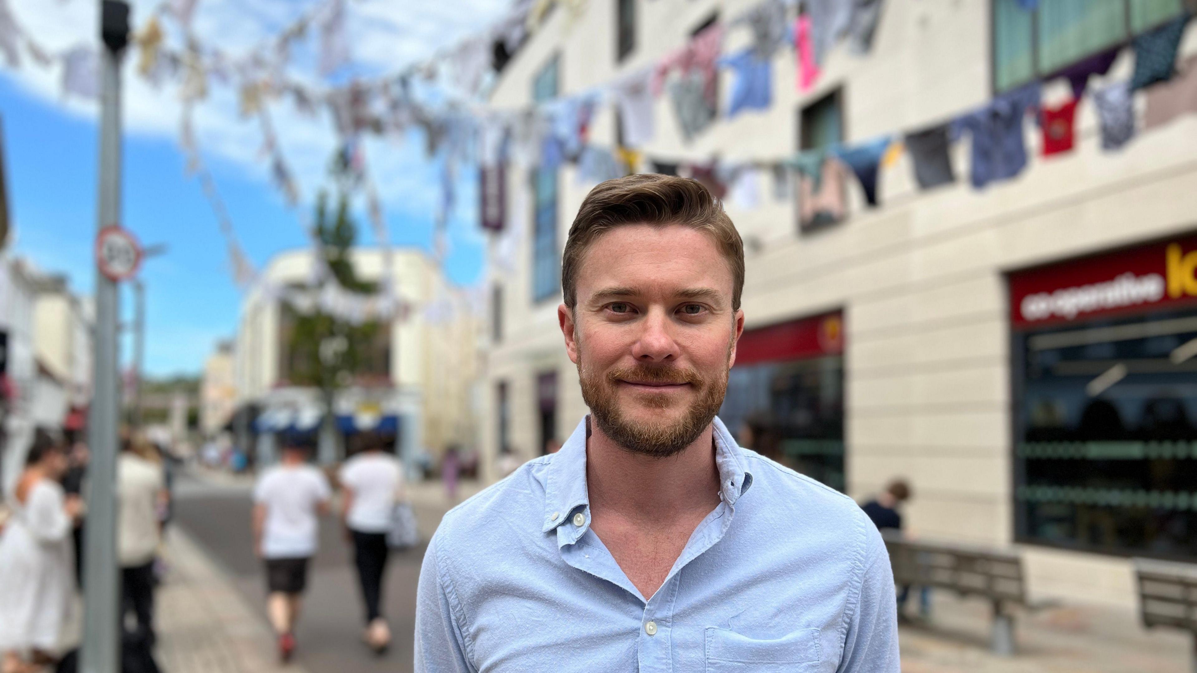 Tom smiles at the camera in the street with the shirts hanging behind him