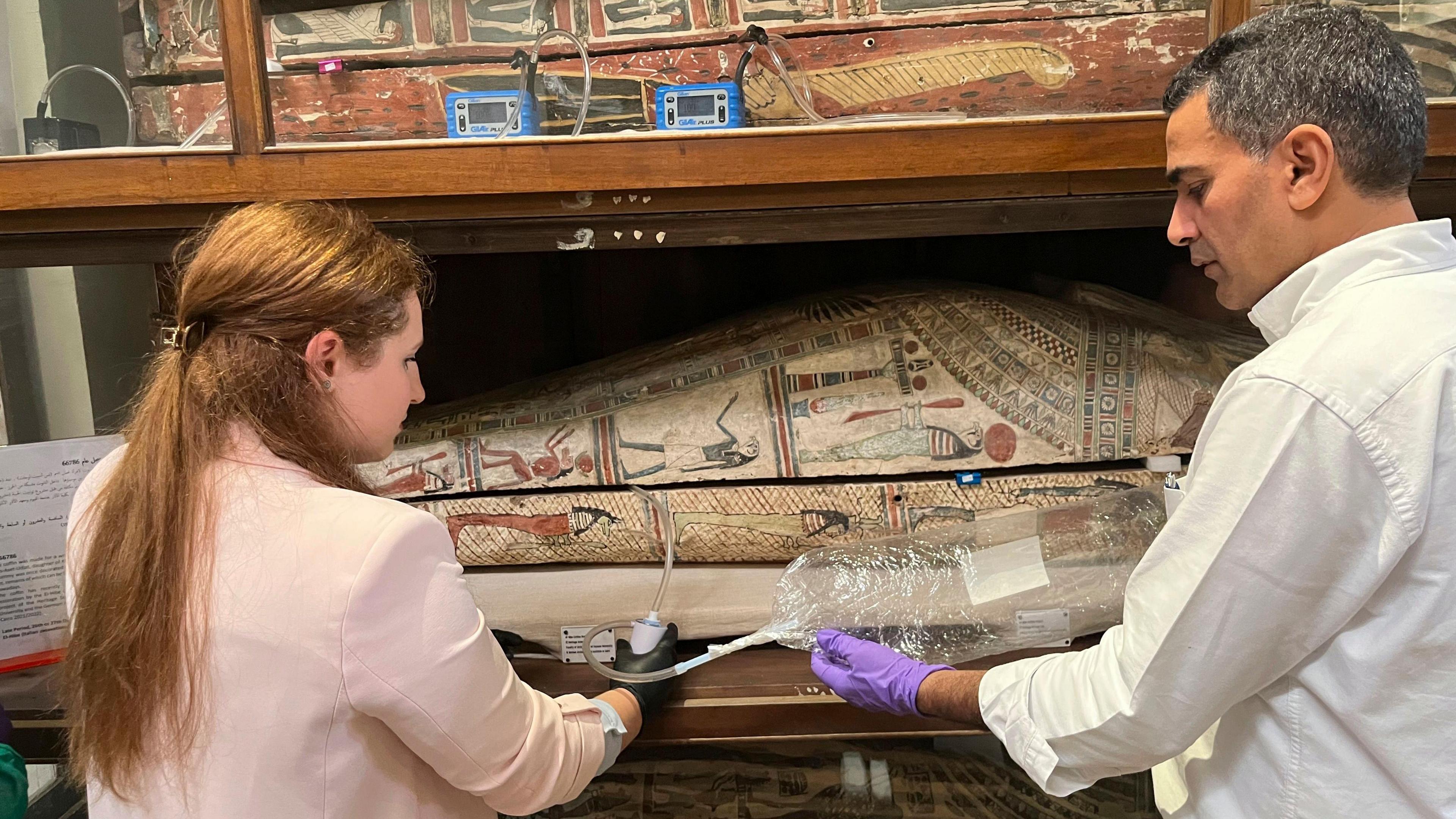 Two researches, dressed in white and wearing gloves, hold a tube into a sarcophagus. The tube is feeding into a bag of air. 