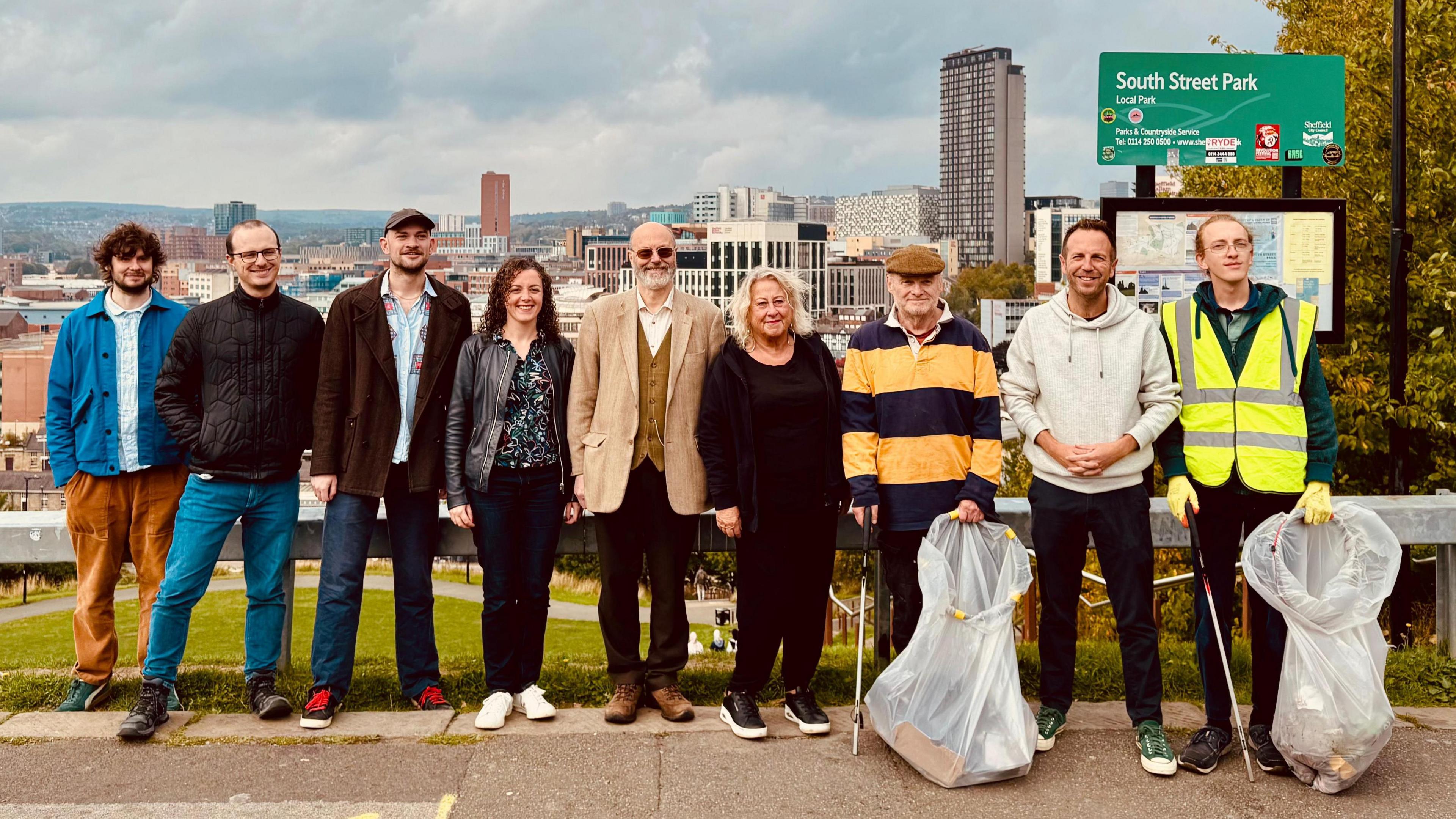 Ten people in a line, some holding bin bags and litter picks. The Sheffield skyline is just about visible behind them, as well as a small park down some steps below.
