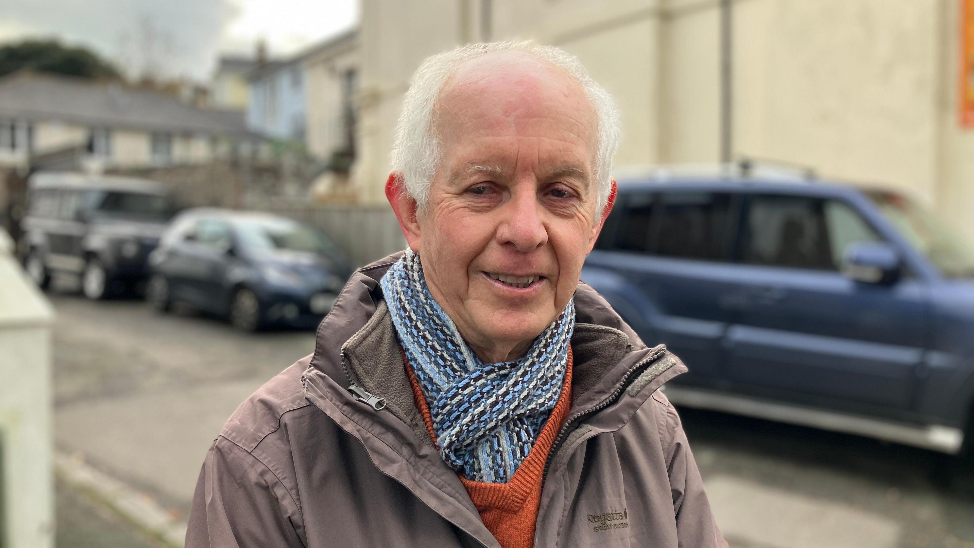 A man stands in a residential street with parked cars in the background. He is wearing a tan coloured coat and blue scarf. 