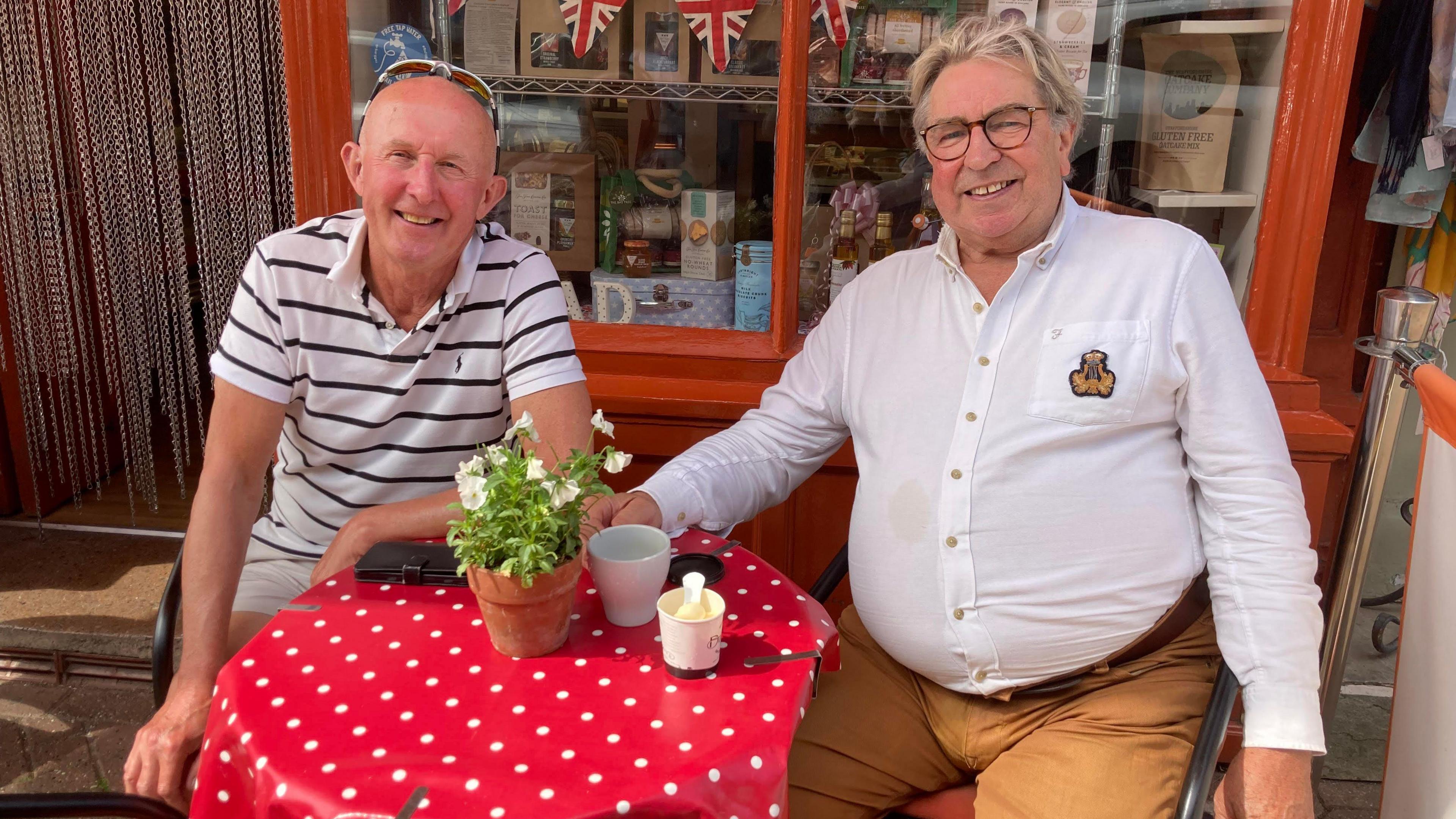 Two men smile while sitting at a small red circular table outside the front of a cafe. A pot plant is on the table along with cups of coffee. The store behind them has an orange frame with historic items in the window.