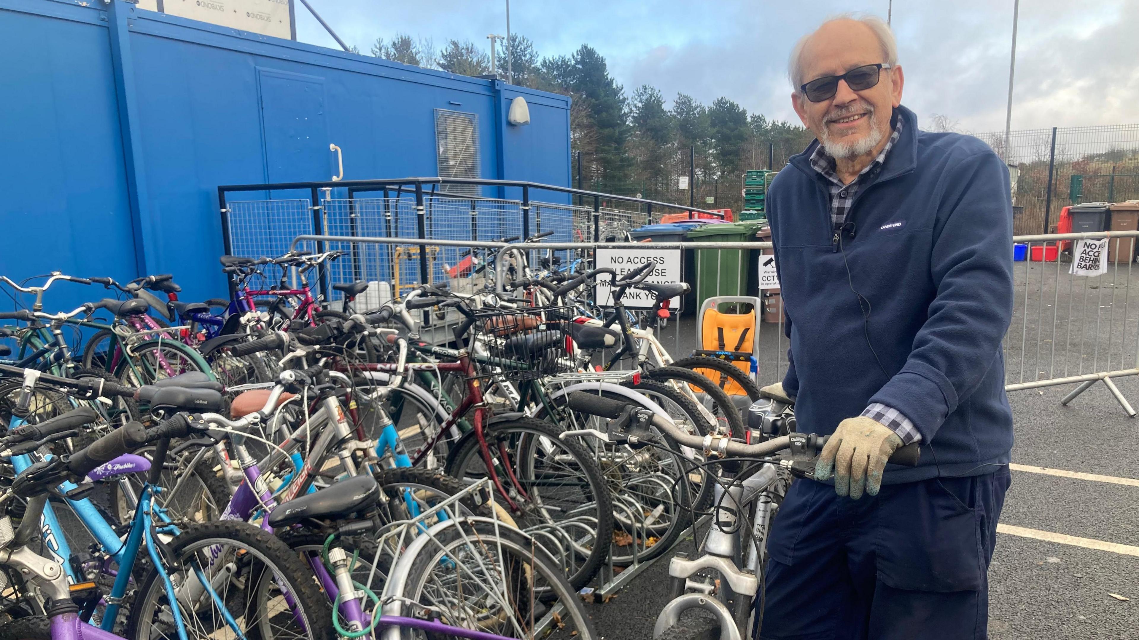 A gentleman with grey hair and a grey beard smiles at the camera while he holds a bike up. He is wearing sunglasses along with a navy jumper, a shirt underneath and gloves over his hands. Tens of bikes are stacked up to the left-hand side of him.